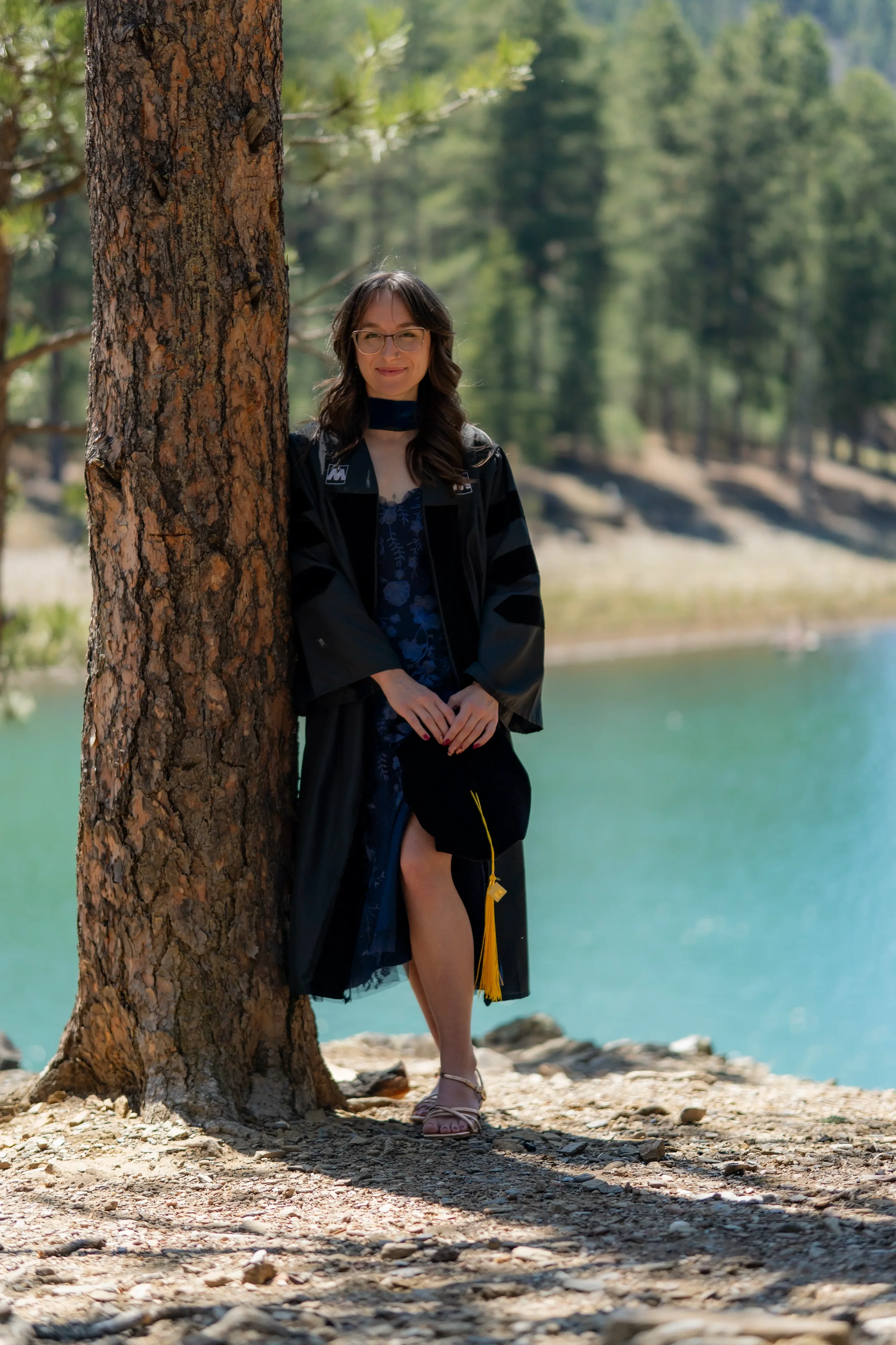 A woman standing next to a tree by a lake, wearing a graduation gown and sash, with one leg partially exposed, in a forested area.