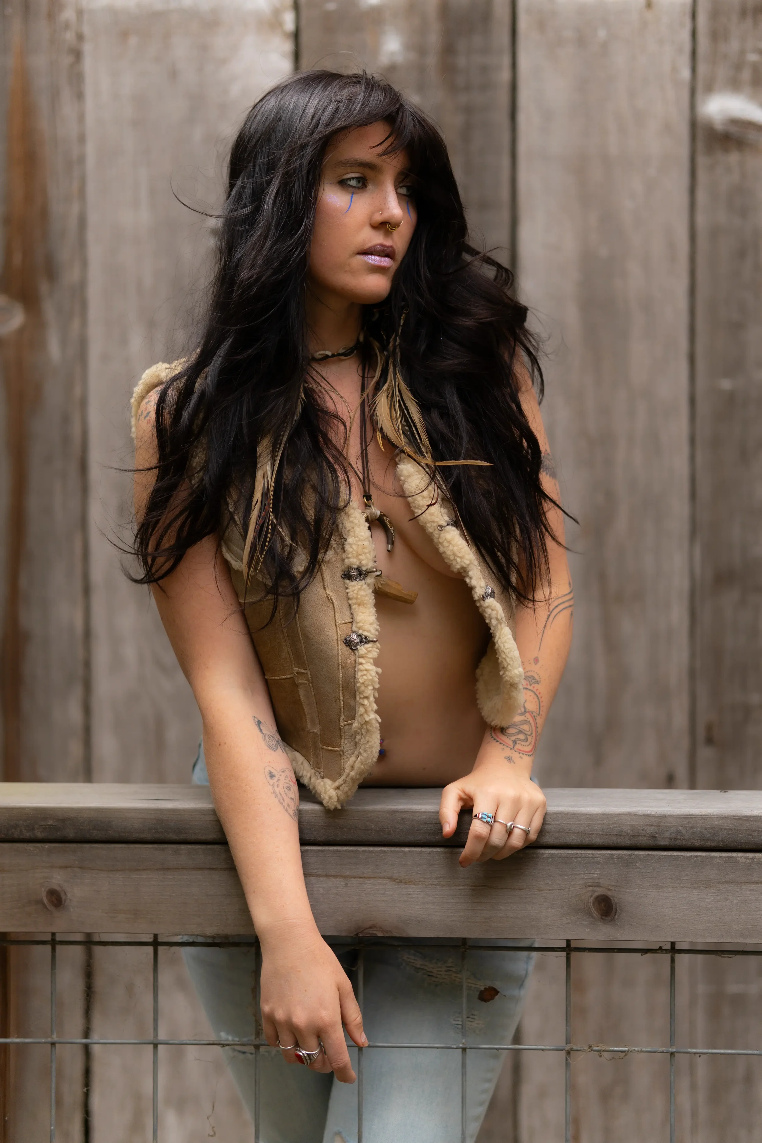 A woman with long black hair and tattoos on her arms is leaning on a wooden railing, wearing a beige vest and light blue jeans, standing in front of a weathered wooden wall.