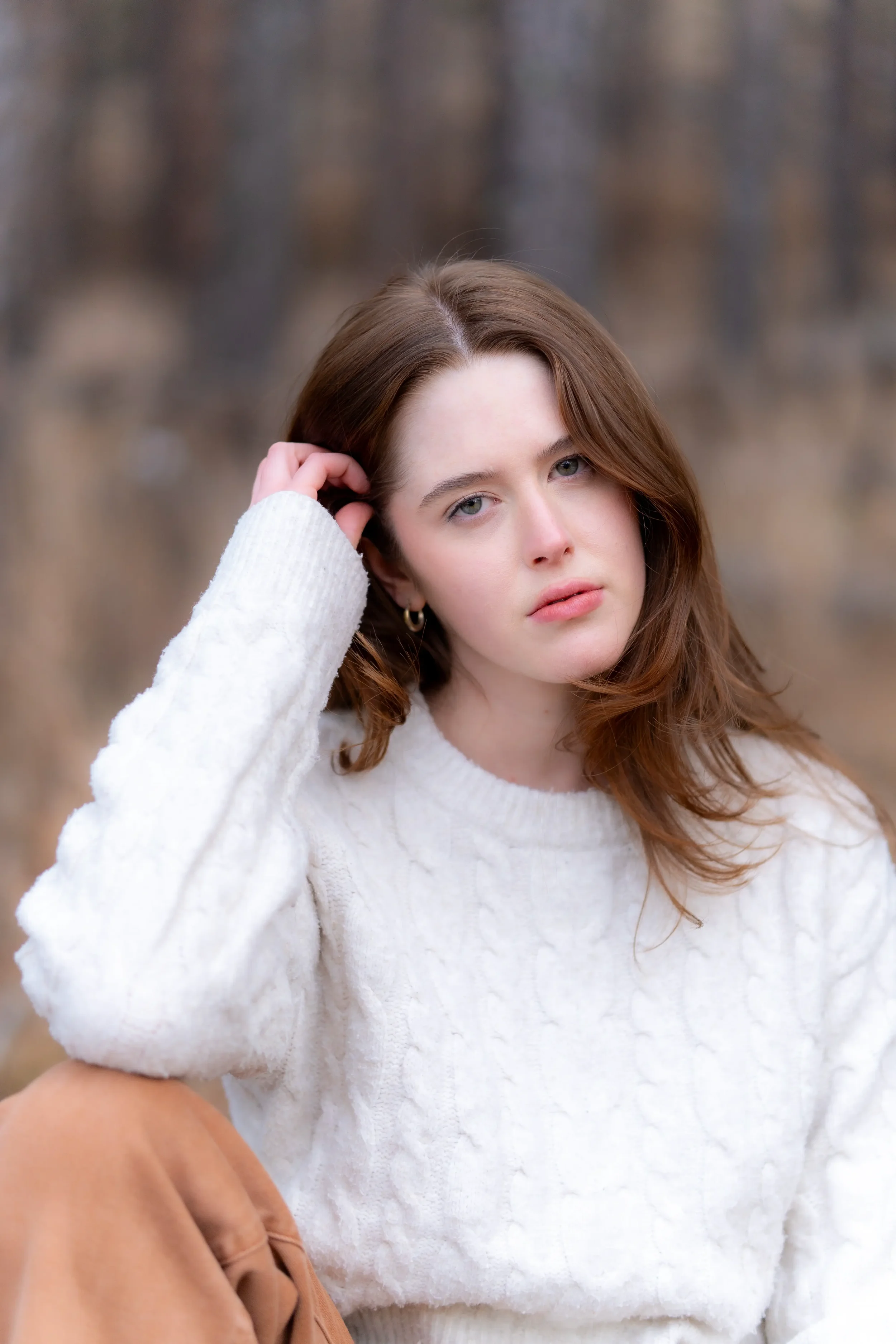 A woman with light skin and long brown hair wearing a white sweater looks into the camera outdoors with a blurred background of trees.