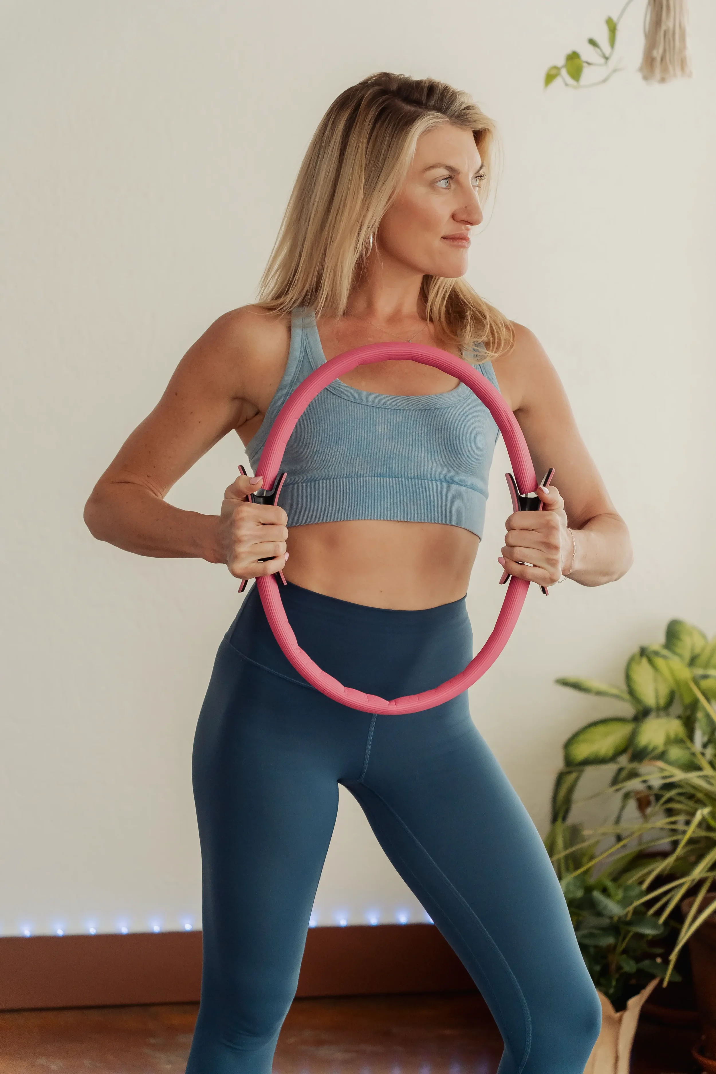 A woman with blonde hair in workout clothes holding a pink exercise ring in front of her waist, standing indoors near a plant.