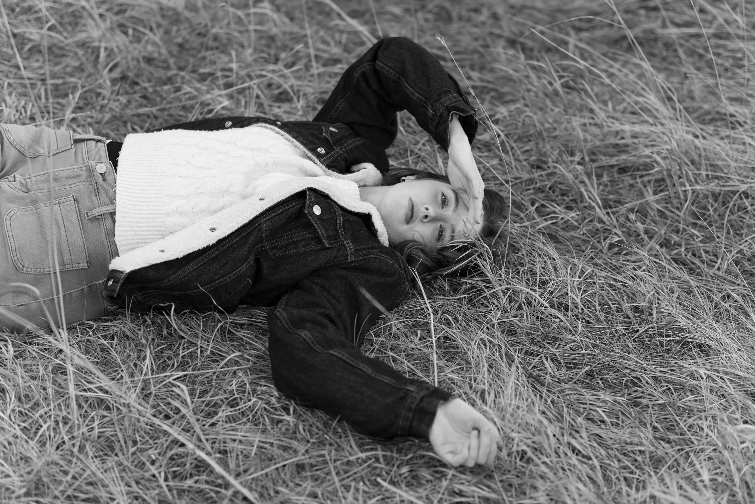 A young woman lying on her back in a field of tall grass, wearing a denim jacket, a white sweater, and jeans, with her hand resting on her forehead and her eyes closed.