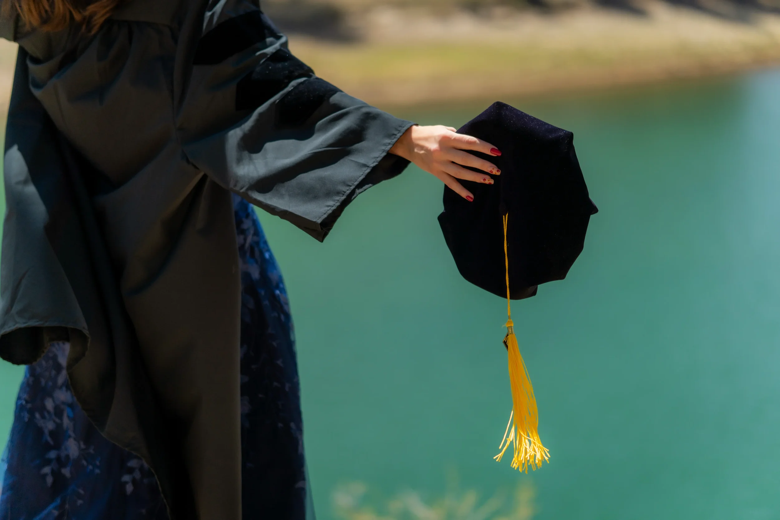 A person in a gray jacket is holding a black graduation cap with a yellow tassel, standing near a body of water with a blurred natural background.