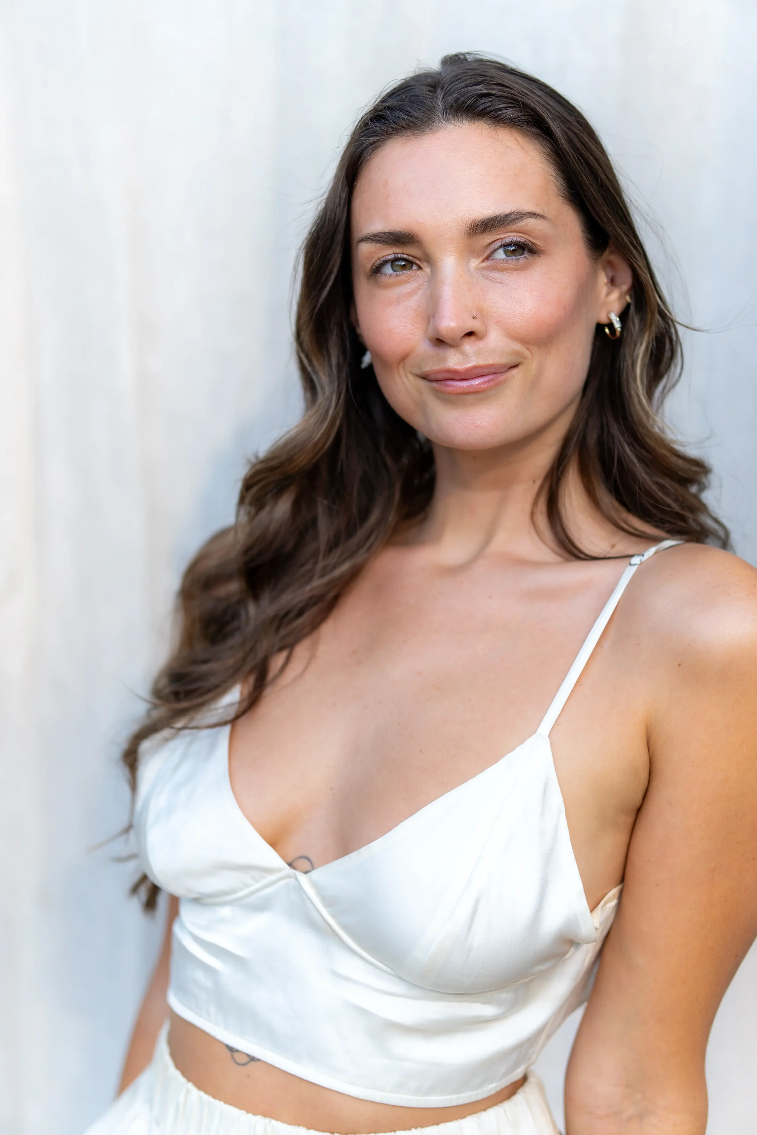 A woman with long wavy brown hair, wearing a white satin crop top with spaghetti straps and matching skirt, standing in front of a light-colored background, smiling softly.