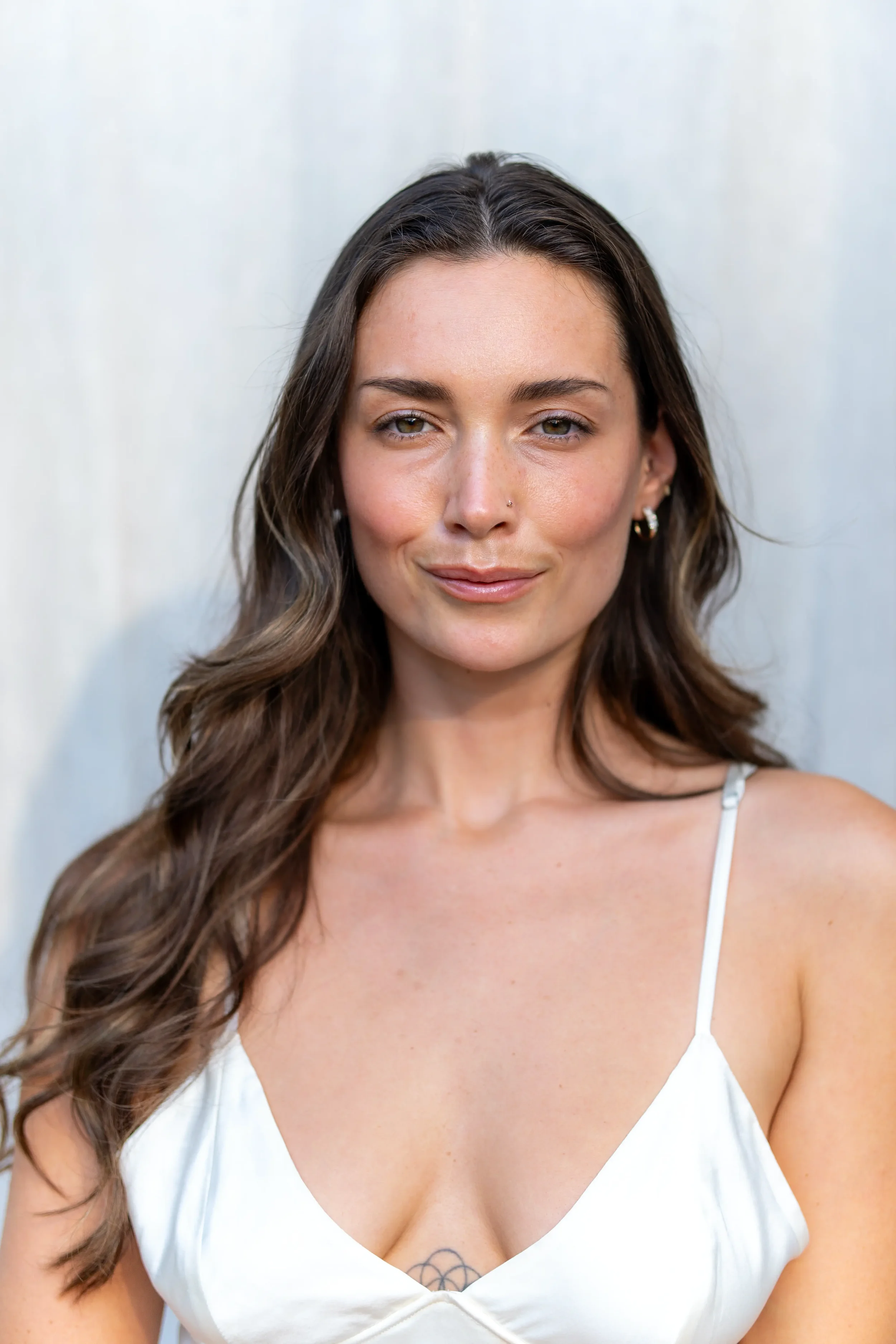 Close-up of a woman with long wavy brown hair, wearing a white top with spaghetti straps, and small hoop earrings, standing against a plain light-colored background.