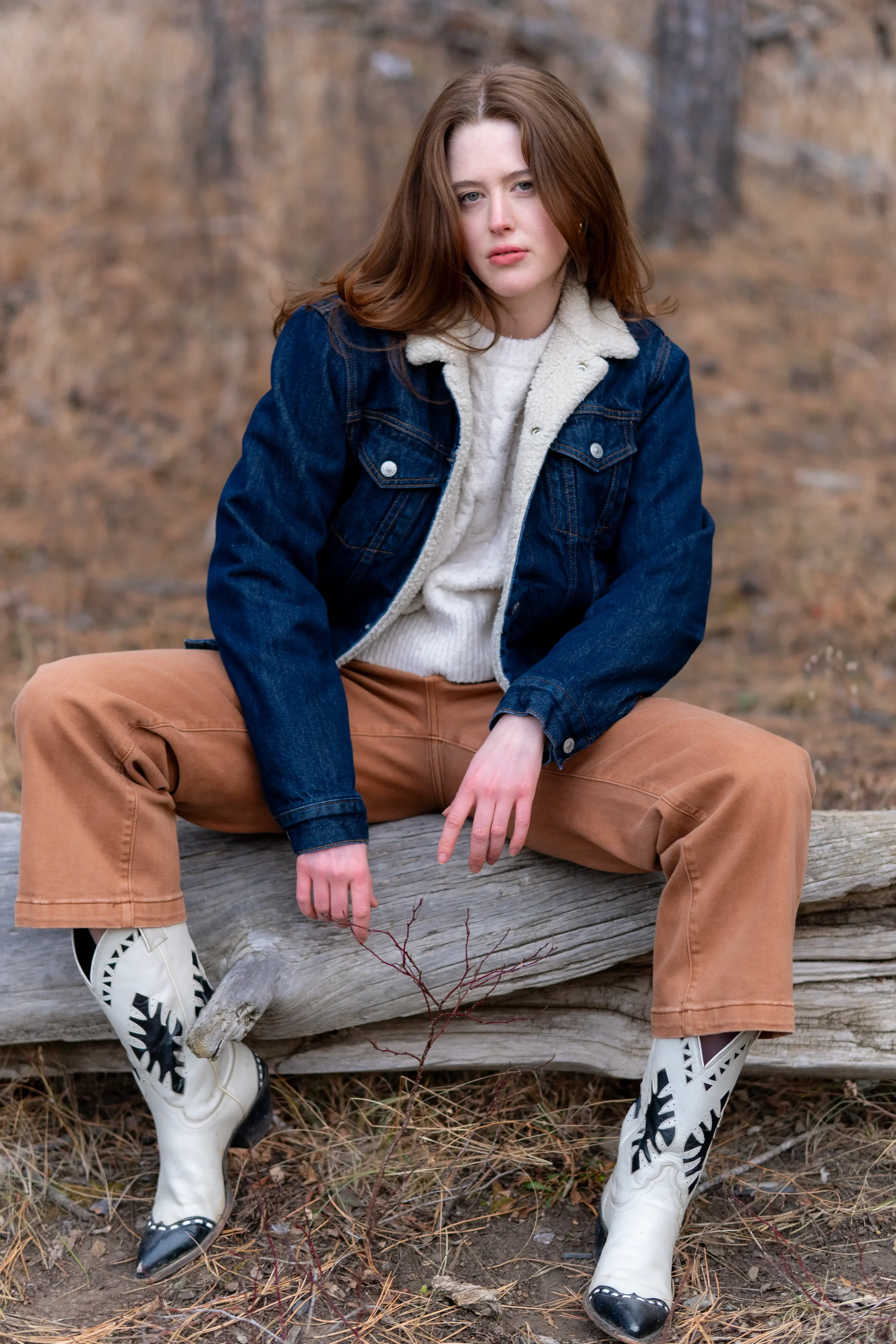 Red-haired woman wearing a denim jacket with shearling lining, cream sweater, beige pants, and white cowboy boots with black accents, sitting on a fallen tree in a natural outdoor setting with brown leaves and twigs.