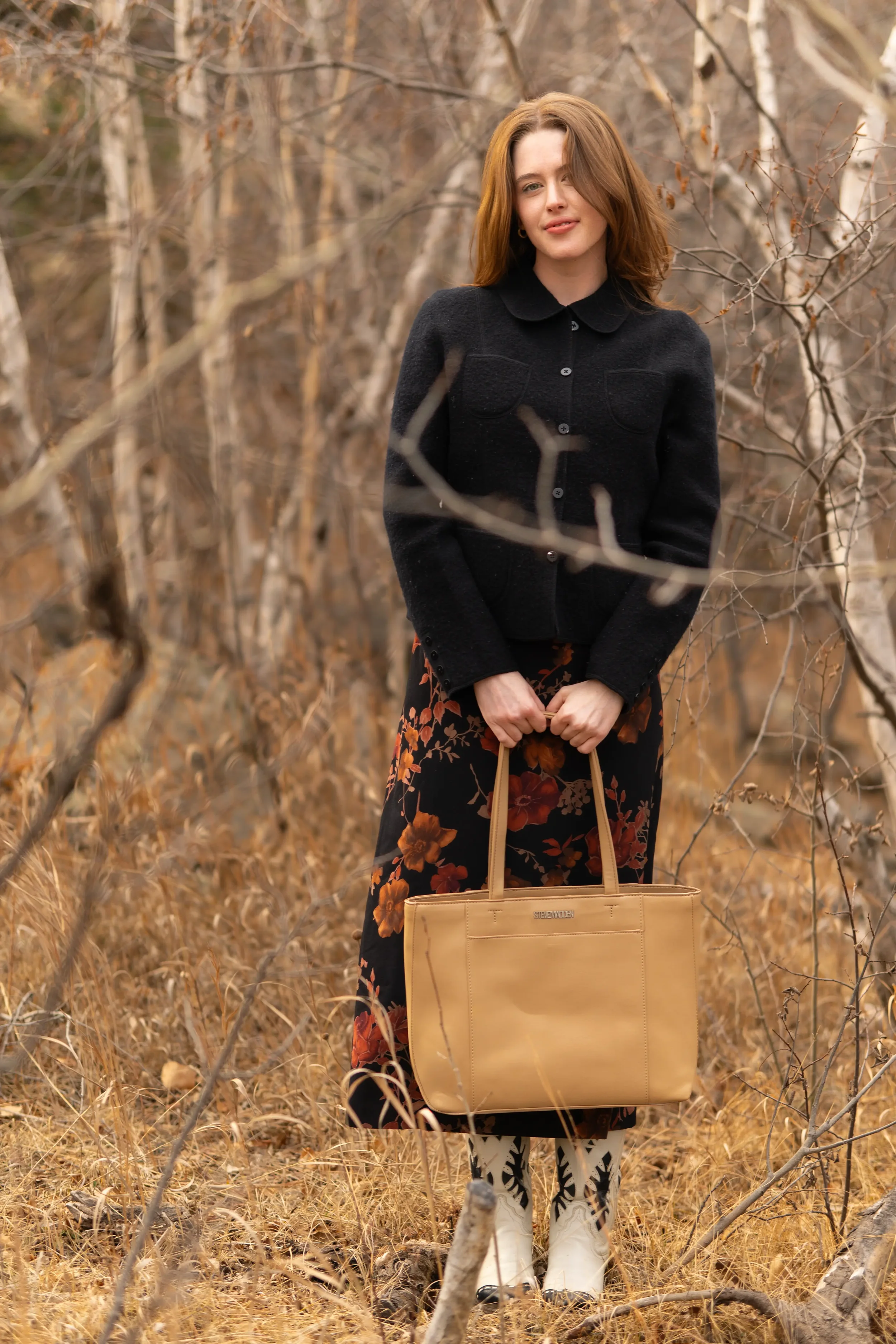A woman with brown hair, wearing a black coat and floral skirt, standing in a dry, leafless woodland area, holding a beige handbag, with a pair of white and black boots.