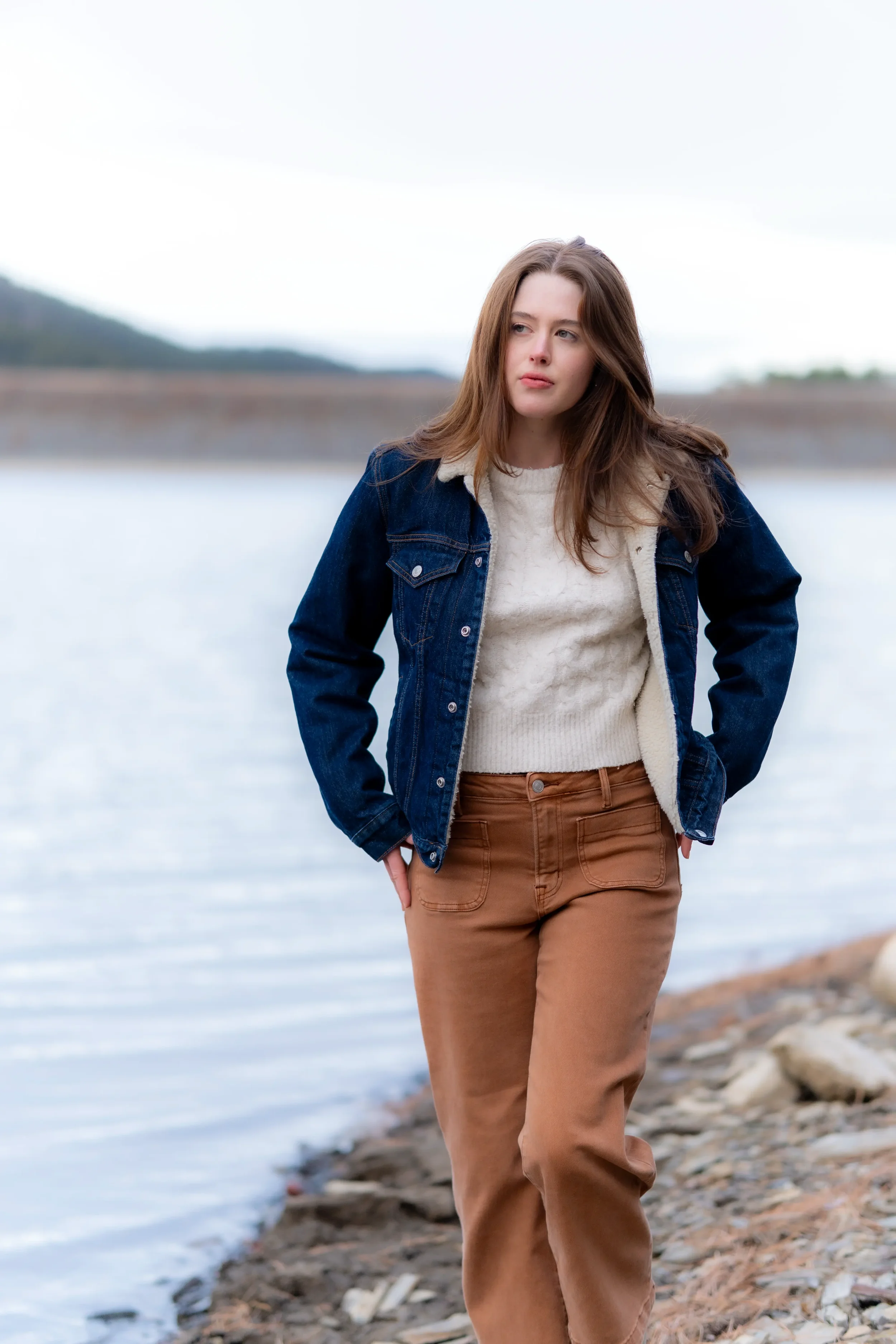 A young woman with long brown hair standing by a body of water on a cloudy day, dressed in a denim jacket, cream sweater, and tan pants.