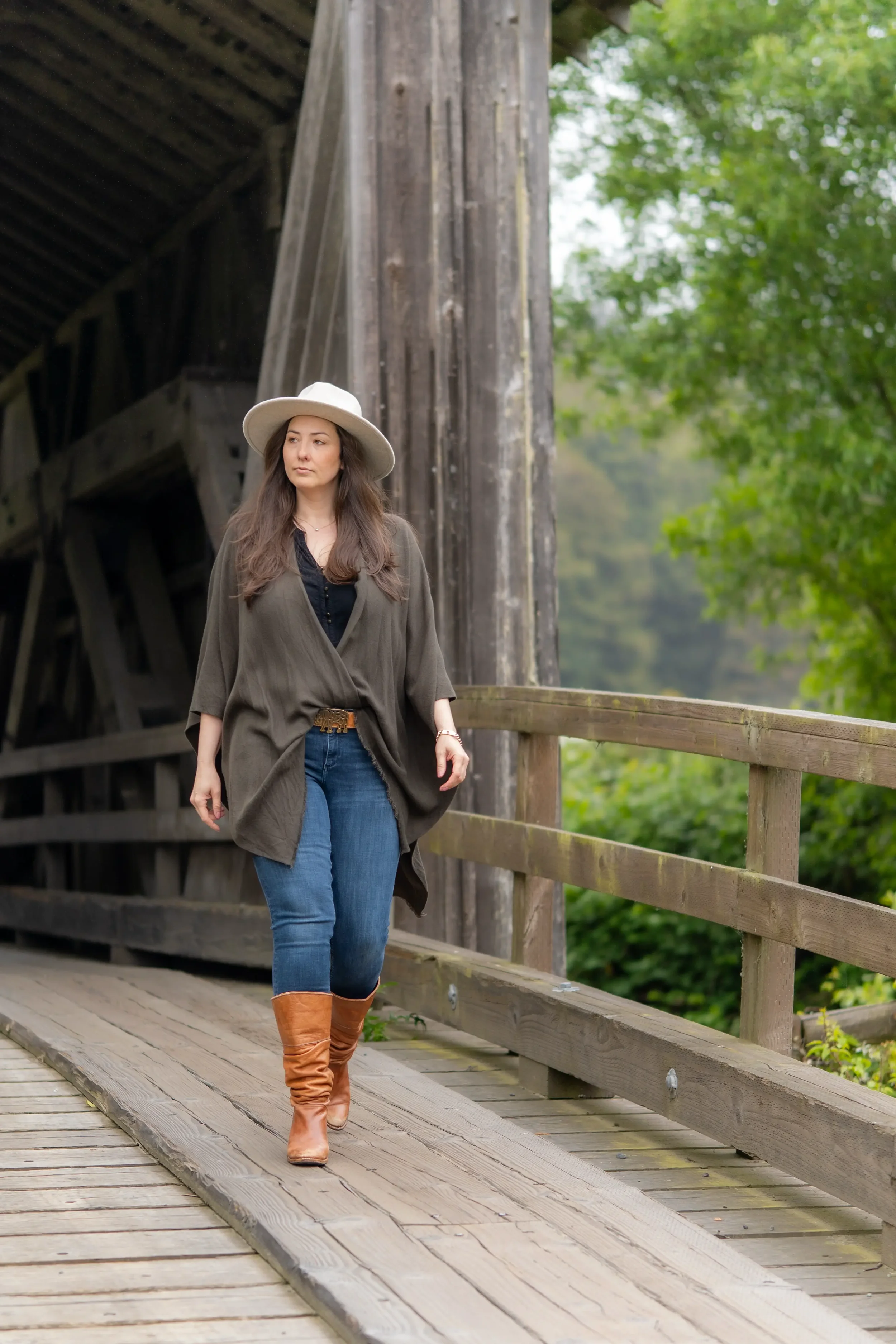 A woman wearing a beige wide-brim hat, an olive green coat, a black top, blue jeans, and tan knee-high boots walking on a wooden bridge outdoors.