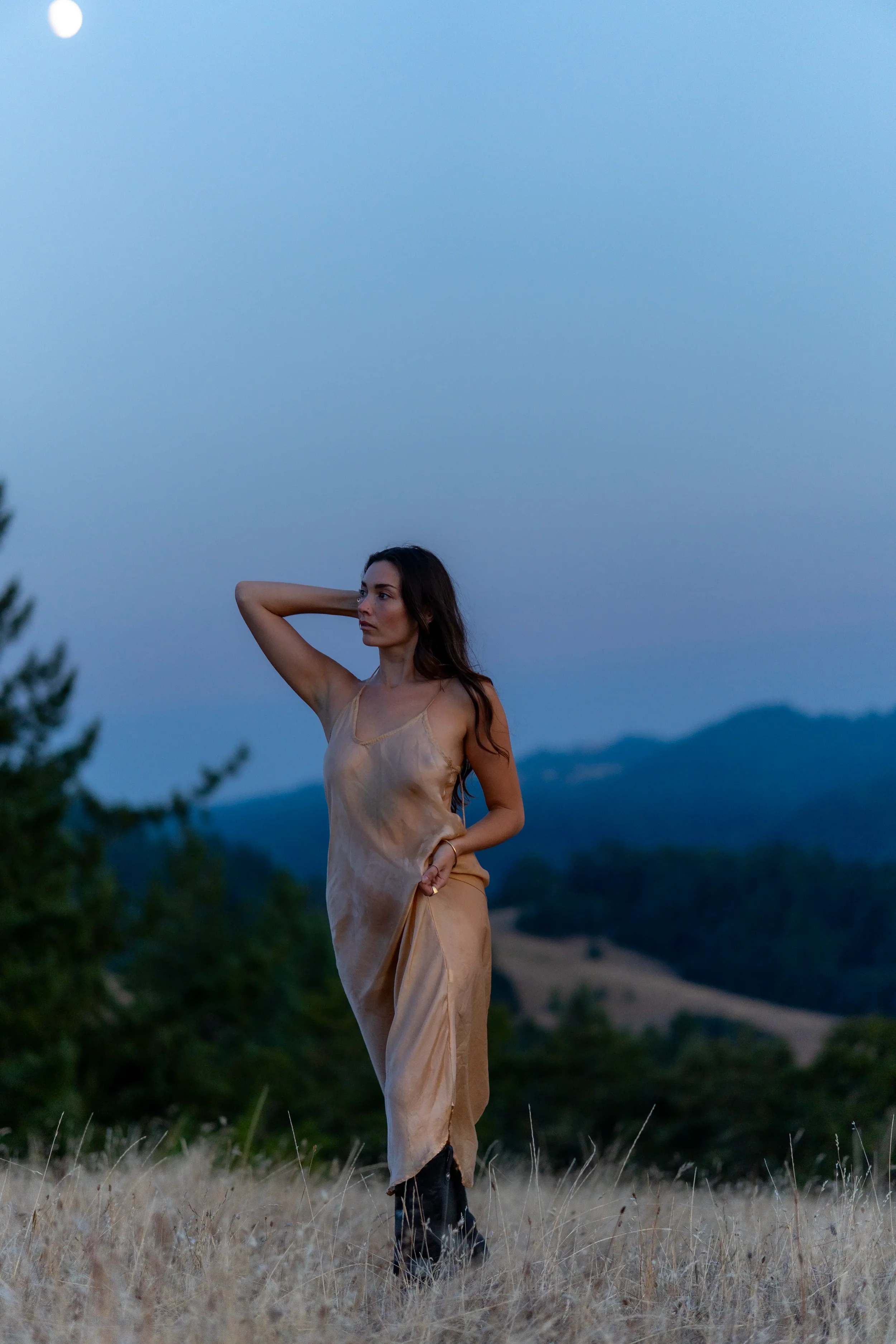 A woman standing in a grassy field with rolling hills and trees in the background during dusk, wearing a tan dress and black boots.