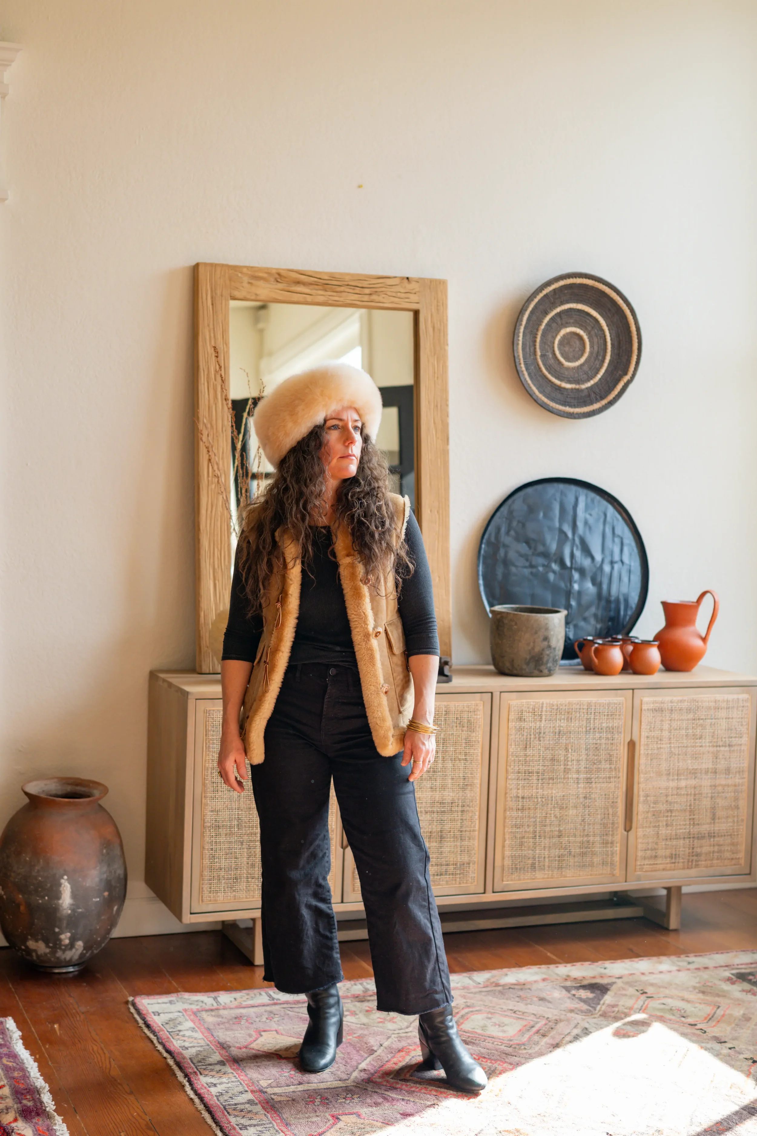 Woman with curly hair wearing a beige furry hat and vest, black long-sleeve shirt, black pants, and black boots standing in a room with a mirror, decorative baskets, pottery, and a patterned rug.