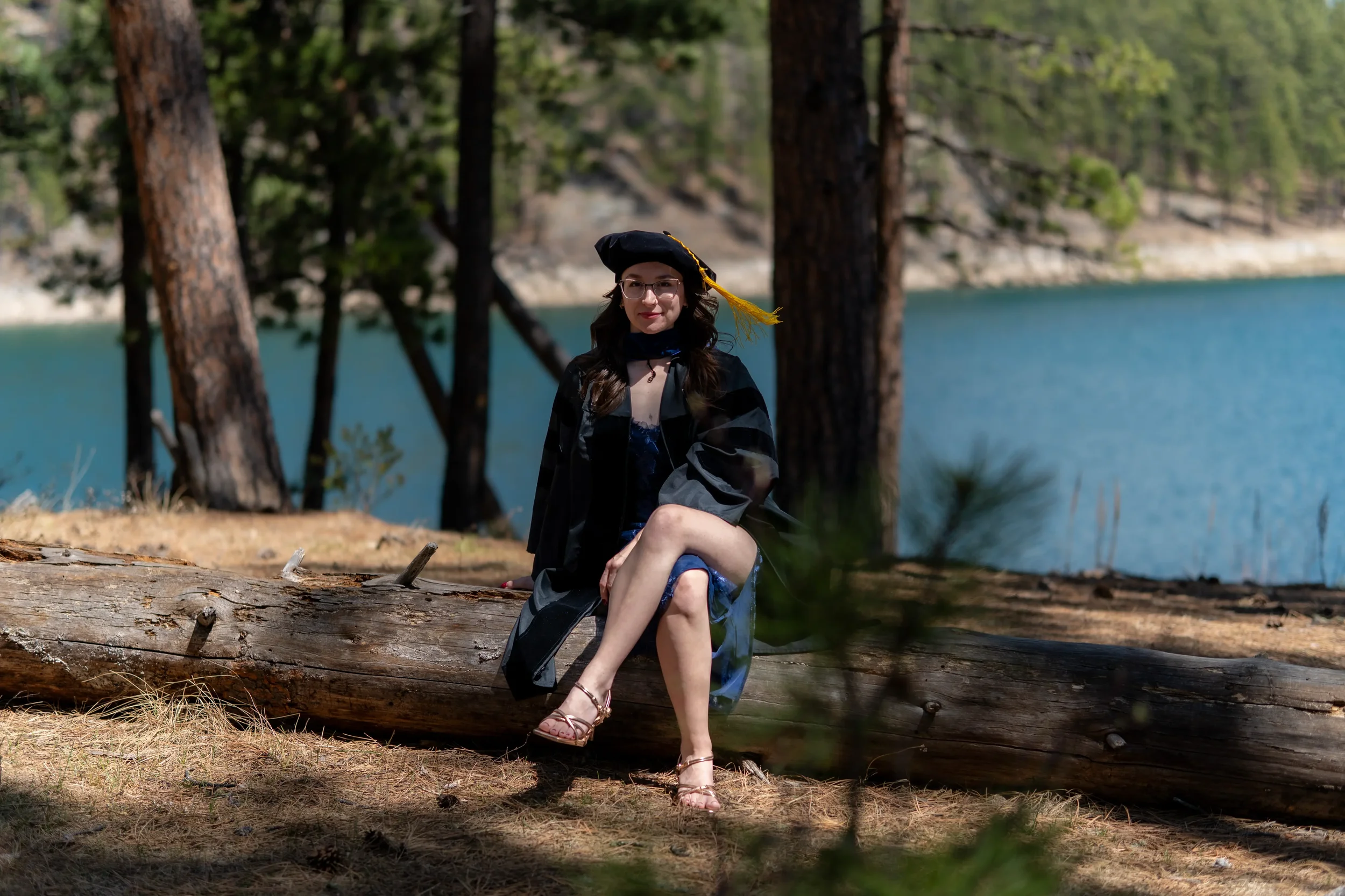 A woman in a graduation cap and gown sitting on a fallen tree trunk near a lake, surrounded by trees, during daytime.
