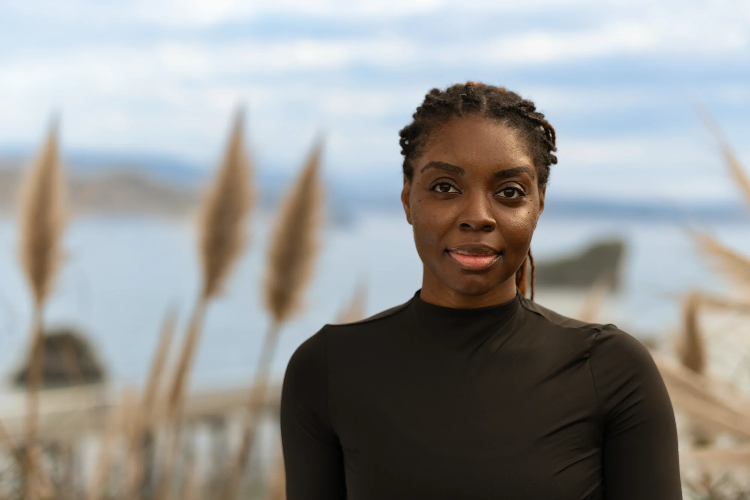 A woman with dark skin and dreadlocks outdoors with blurred grass and trees in the background.