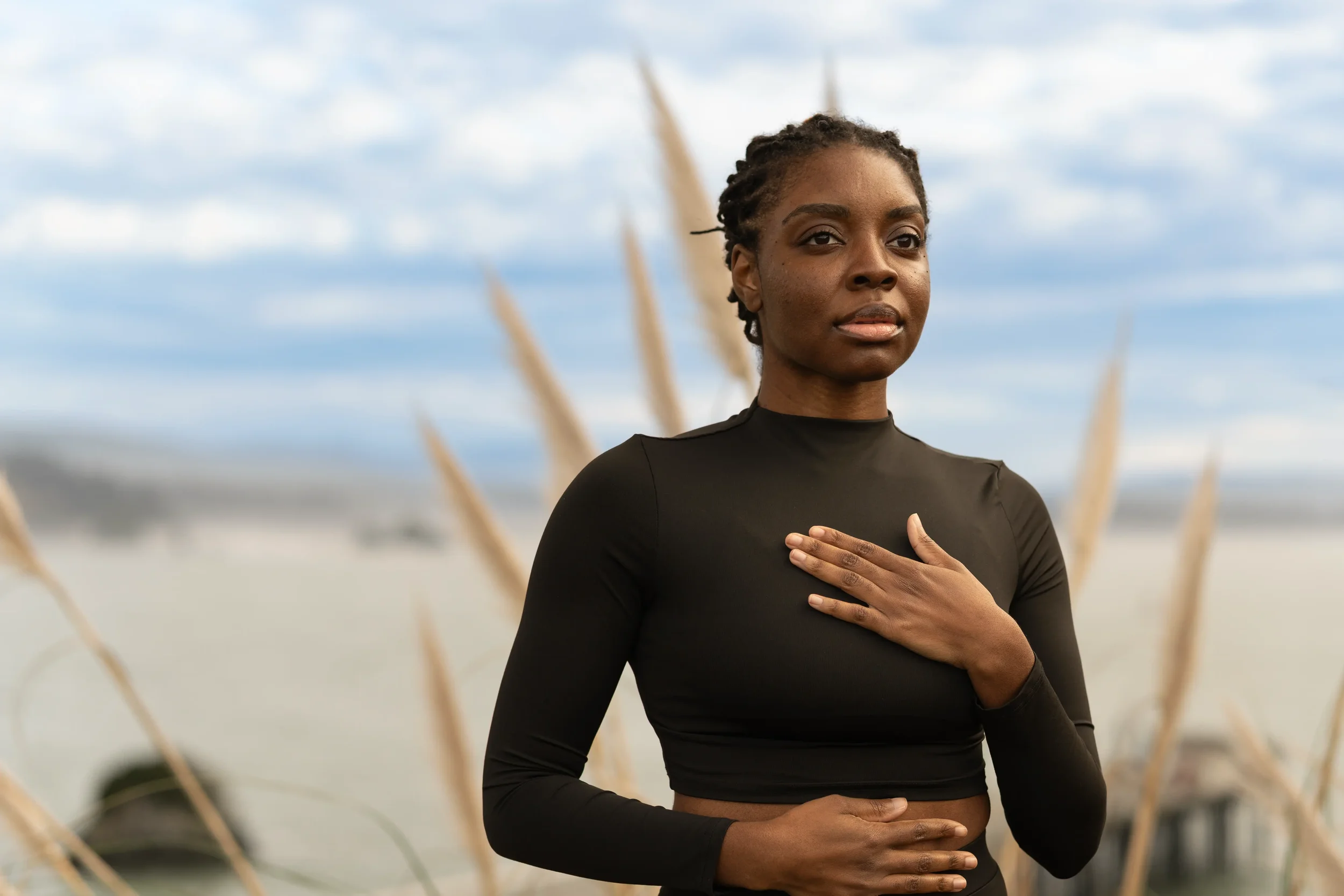 A woman with dark skin and dreadlocks standing outdoors in a black long-sleeve top with her right hand on her chest, looking thoughtfully into the distance, with tall grass and a cloudy sky in the background.