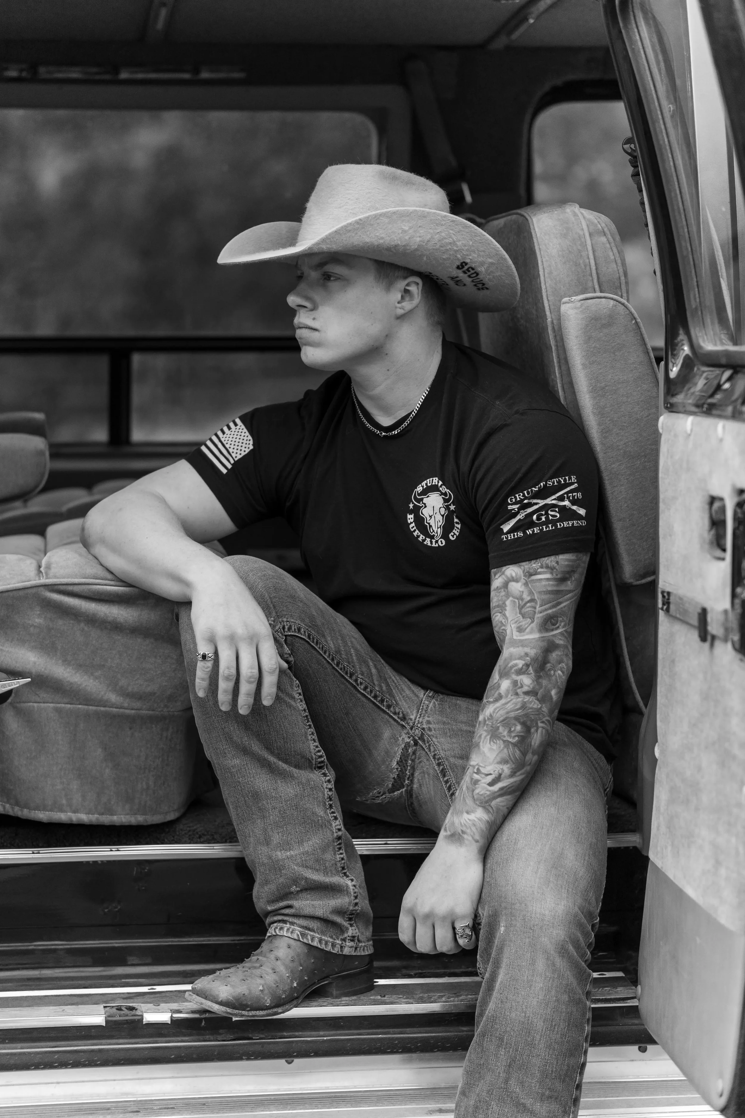 A young man wearing a cowboy hat, black t-shirt with Buffalo Bills logo, and jeans, sitting inside a vehicle with an open door. He has tattoos on his arm and is looking pensively to the side.