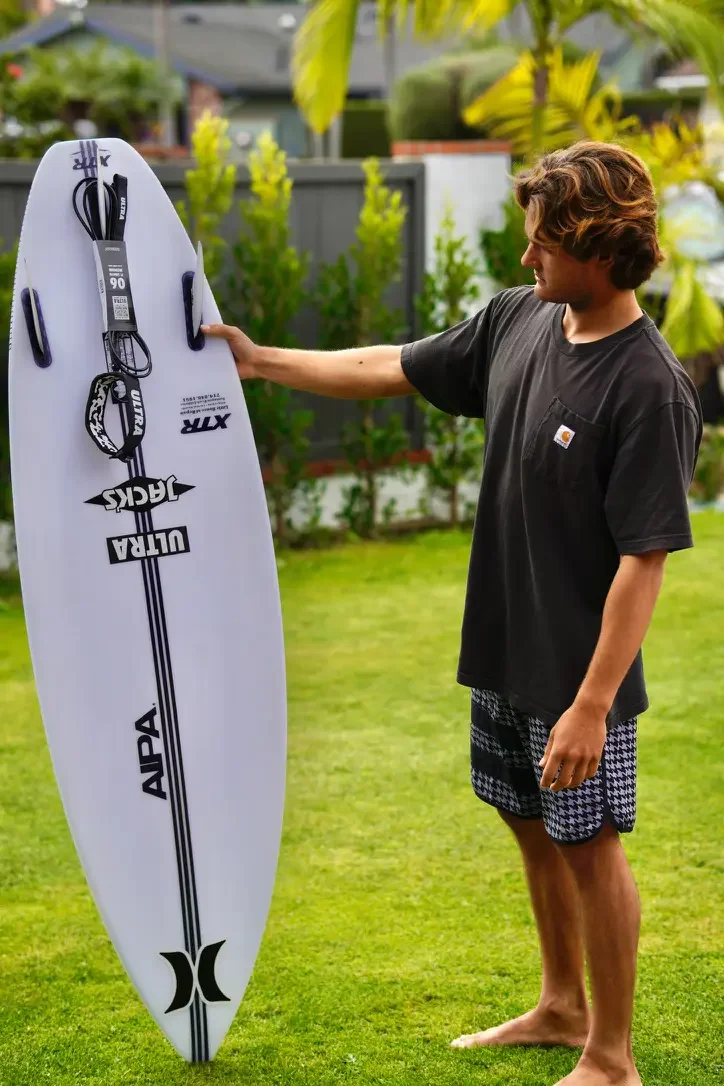 A young man standing barefoot on grass and holding a white surfboard with black and white stickers in a backyard with trees and houses in the background.