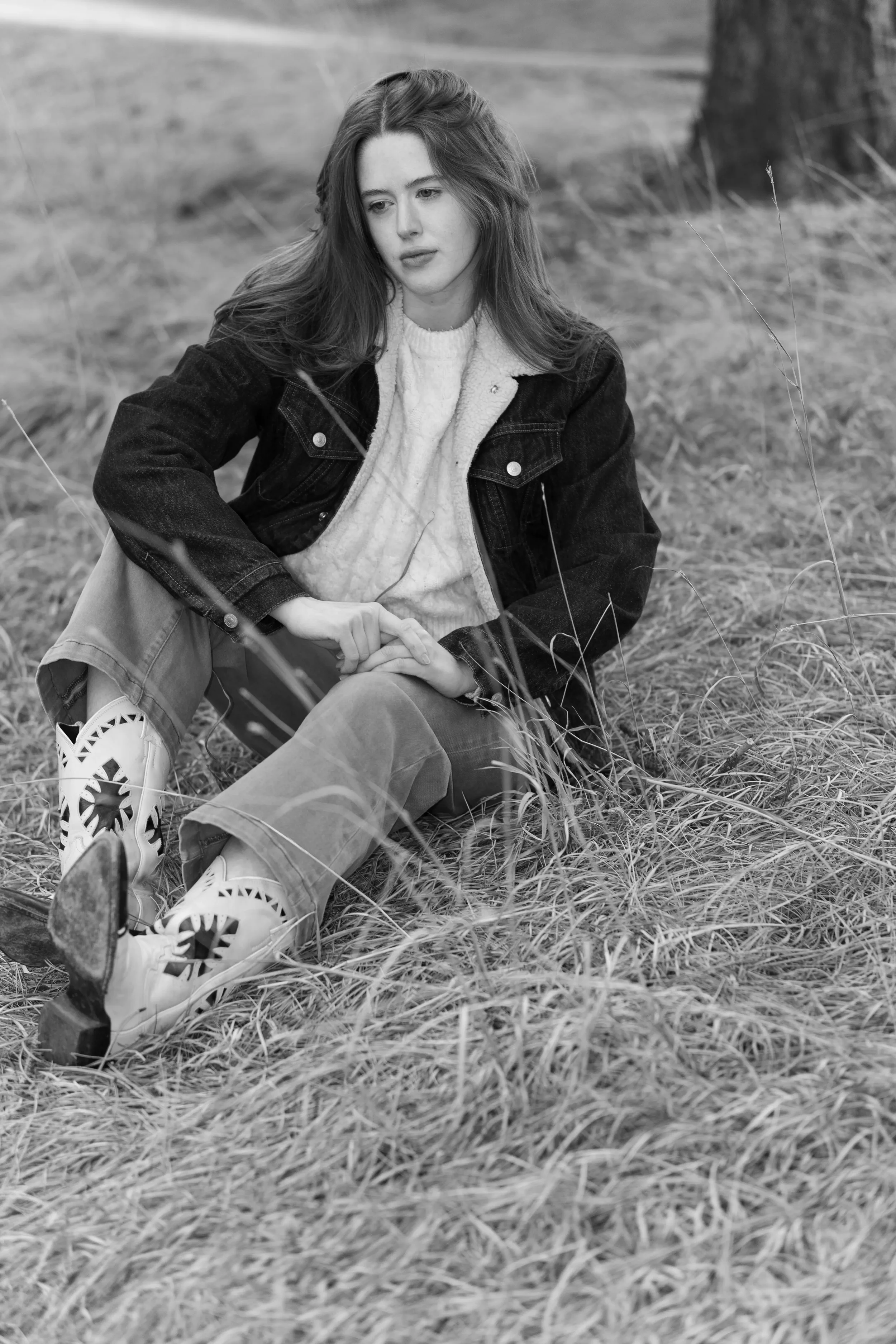 A young woman with long hair sitting on the grassy ground outdoors, wearing a denim jacket, sweater, and patterned boots.