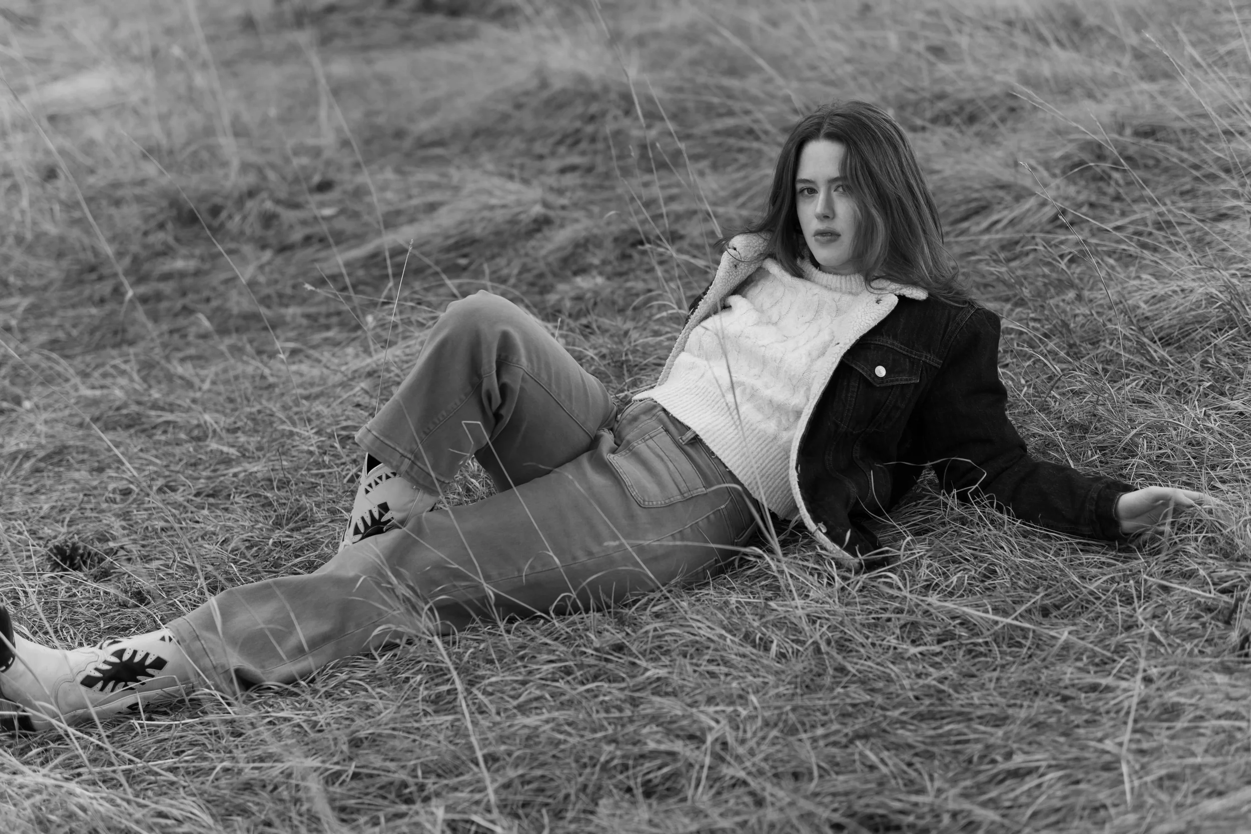 A woman with long hair lying on grass in an open outdoor area, wearing a sweater, jeans, and a jacket, with a serious expression.