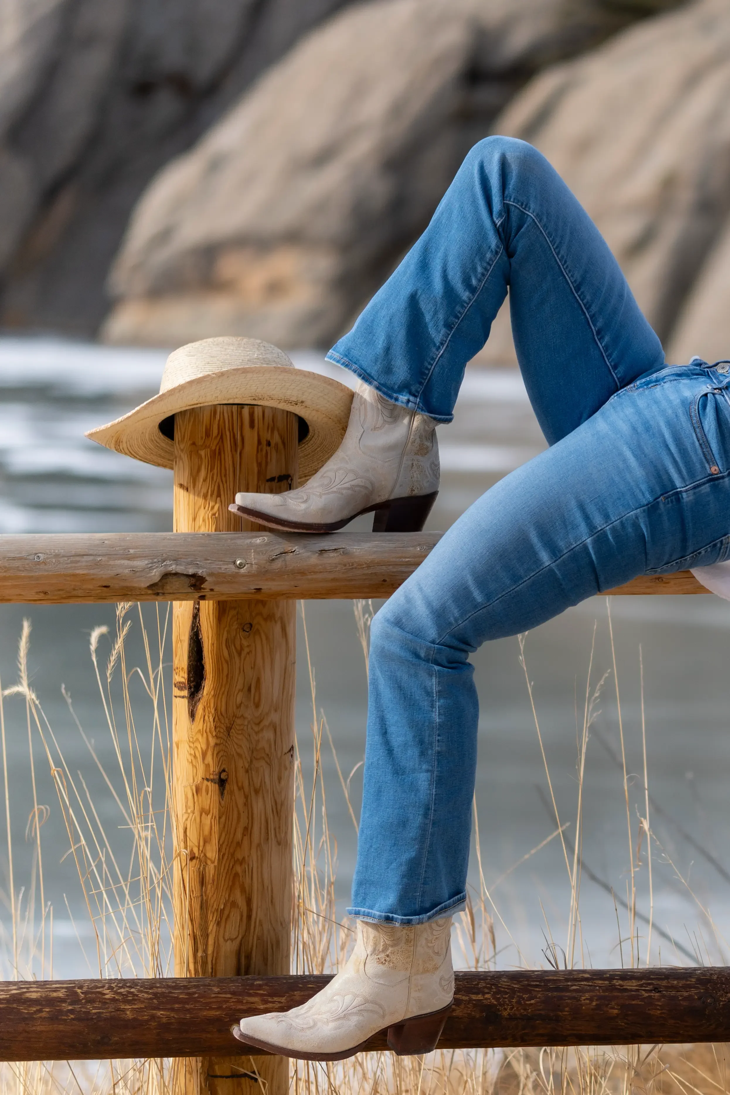 Close-up of a person leaning against a wooden railing by the water, wearing blue jeans, white cowboy boots, and a cowboy hat resting on the railing, with rocky shoreline and ocean in the background.