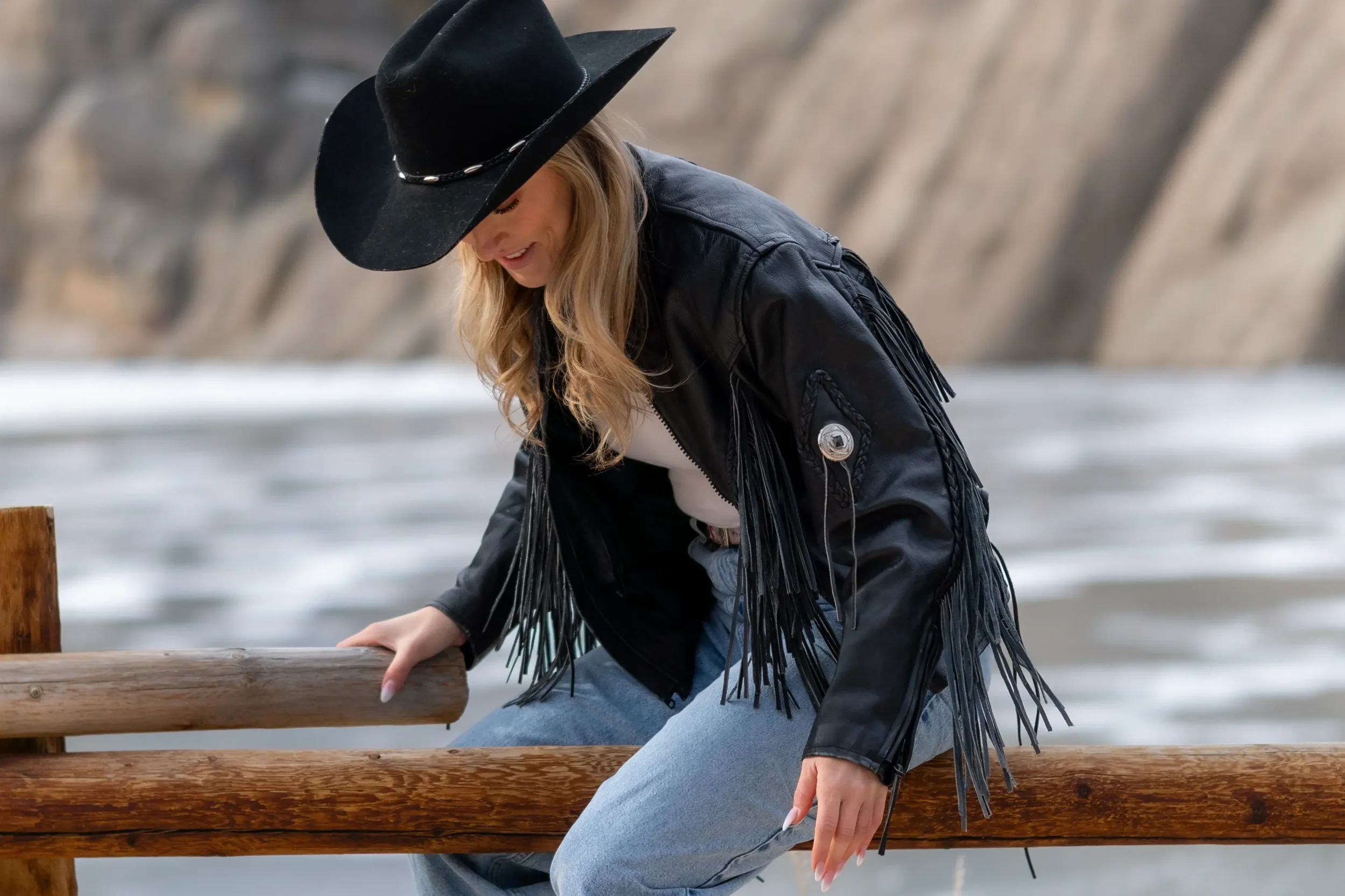 A woman wearing a black cowboy hat, black fringe leather jacket, and gray jeans sitting on a wooden bench near water, looking down.