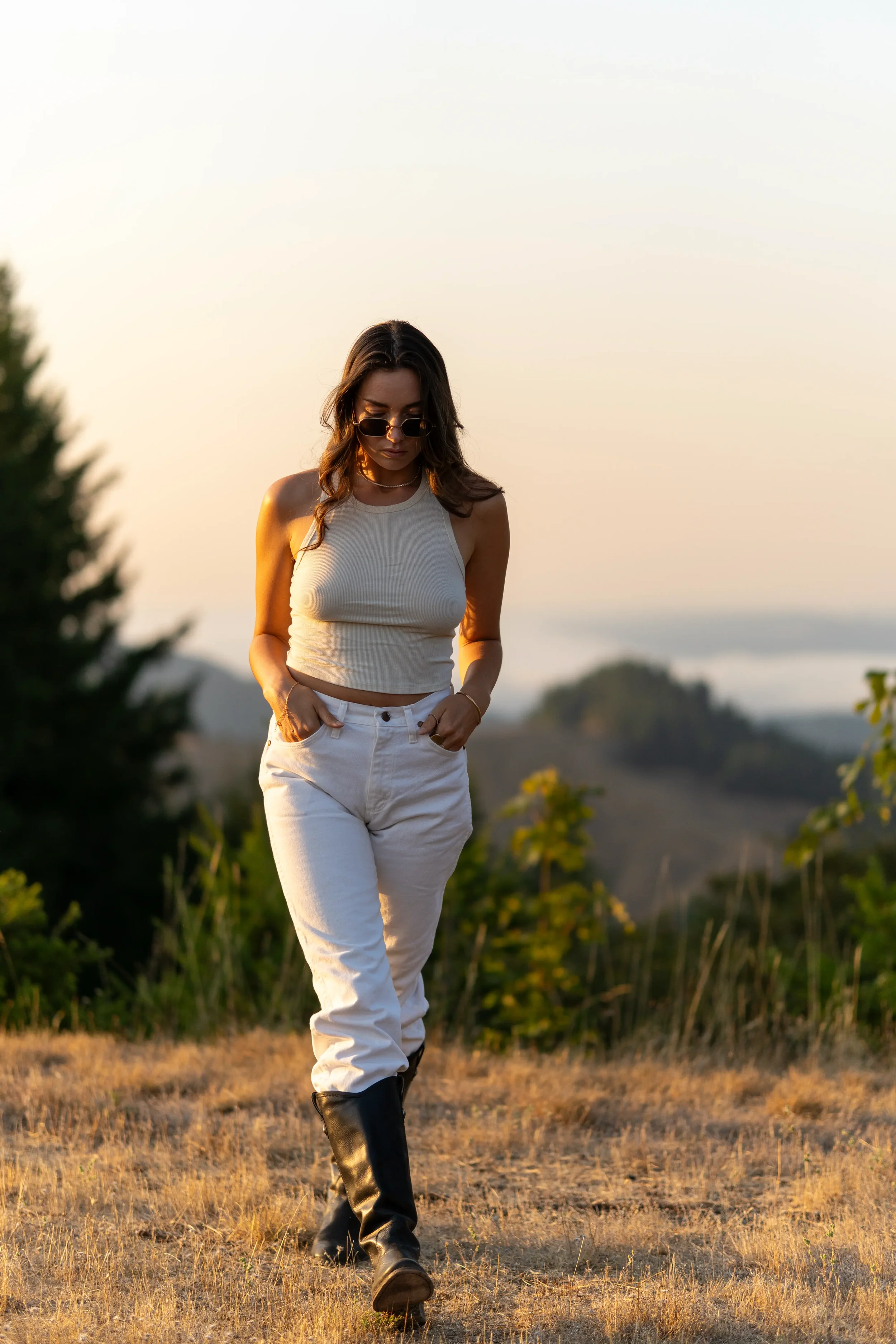 A young woman with wavy brown hair wearing sunglasses, a white tank top, white pants, and black boots walking in a field during sunset.