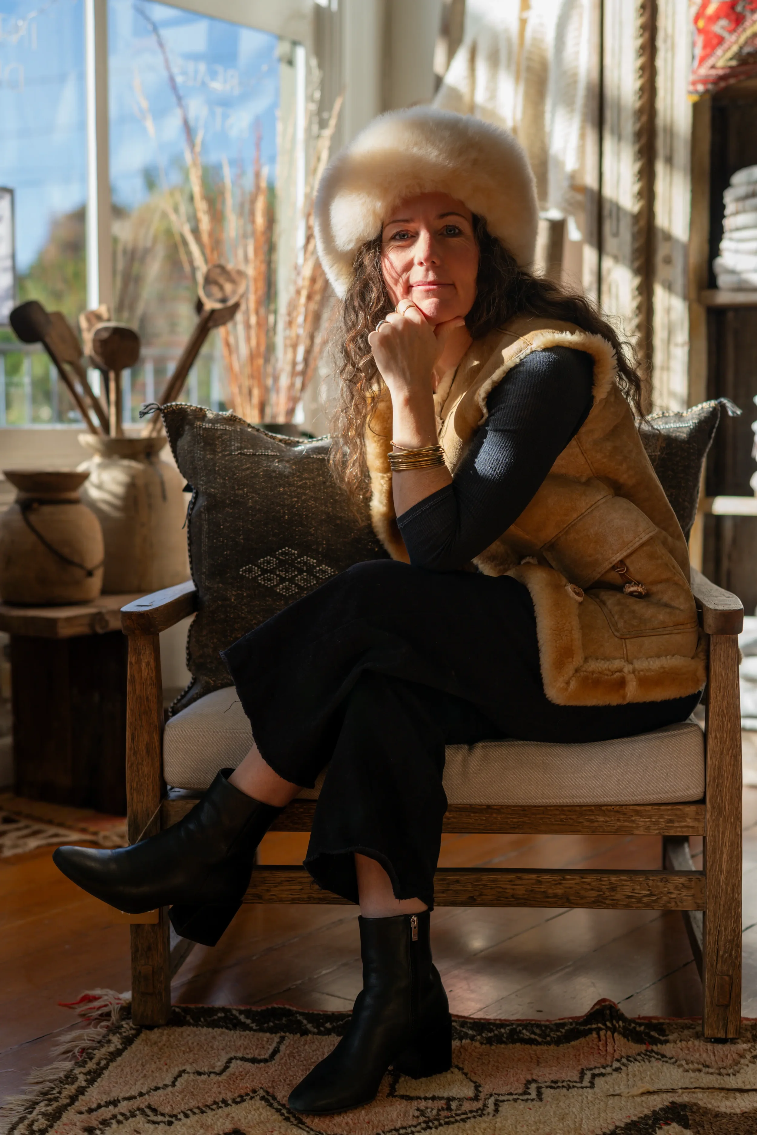 A woman sitting on a wooden bench inside a cozy room with sunlight streaming through a window. She wears a large fluffy hat, a black long-sleeve top, a tan fur vest, black pants, and black boots. She rests her chin on her hand and looks thoughtfully 