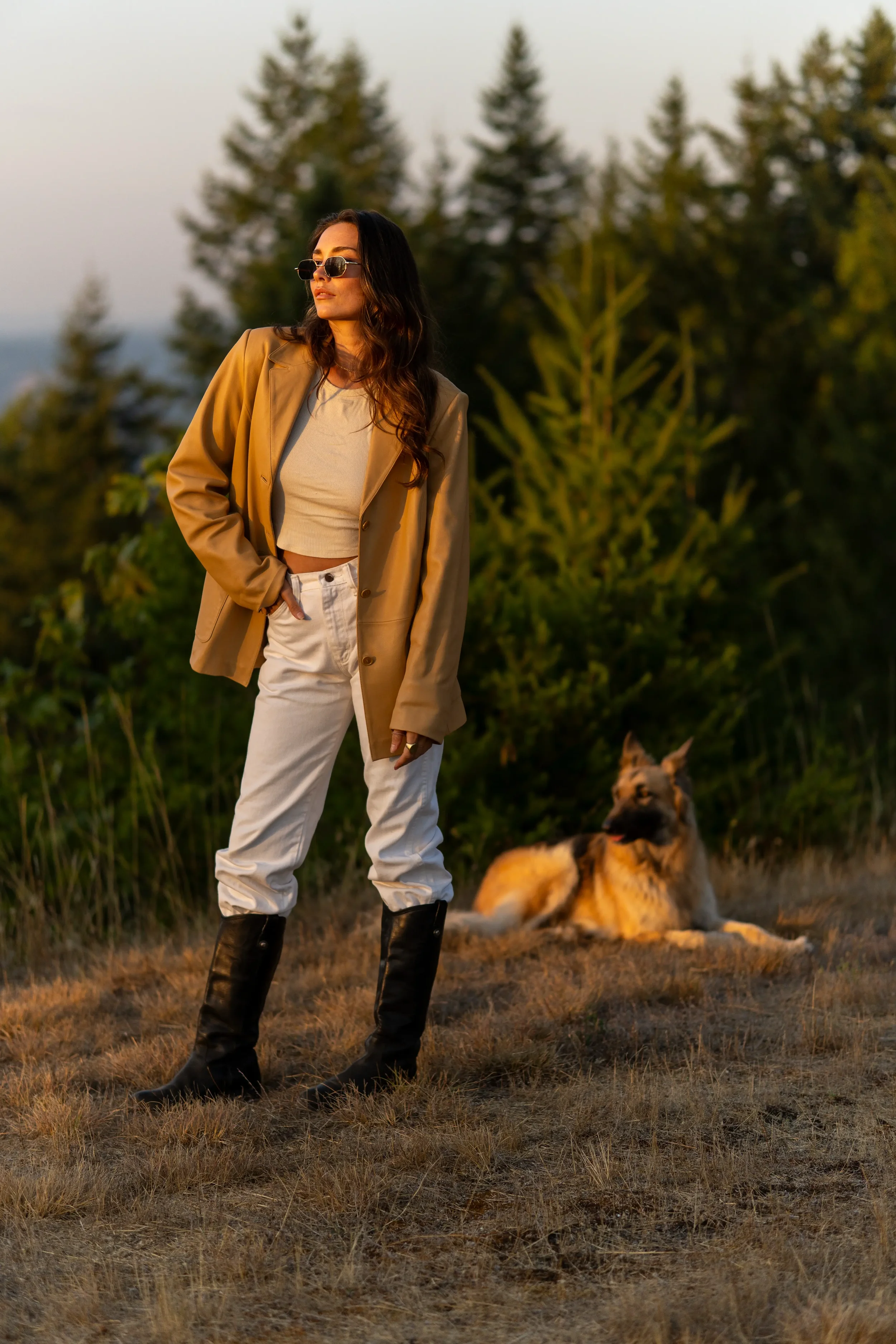 A woman with long wavy hair wearing sunglasses, a tan blazer, cream crop top, white pants, and black boots standing outdoors at sunset, with a large dog lying on the ground nearby and trees in the background.