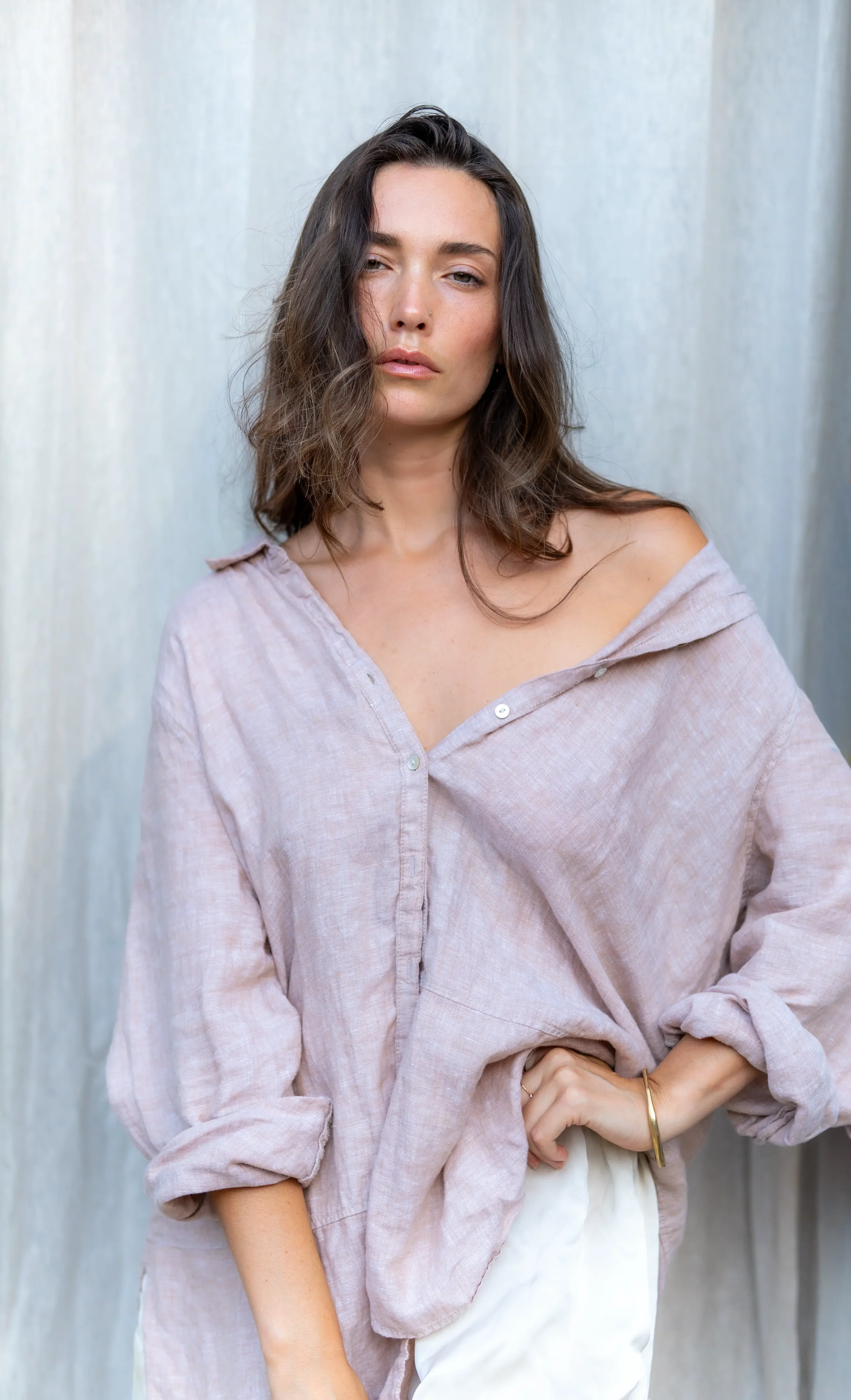 A woman with wavy dark brown hair and light makeup wearing a loose beige button-up shirt and white pants, standing against a plain, light-colored backdrop.