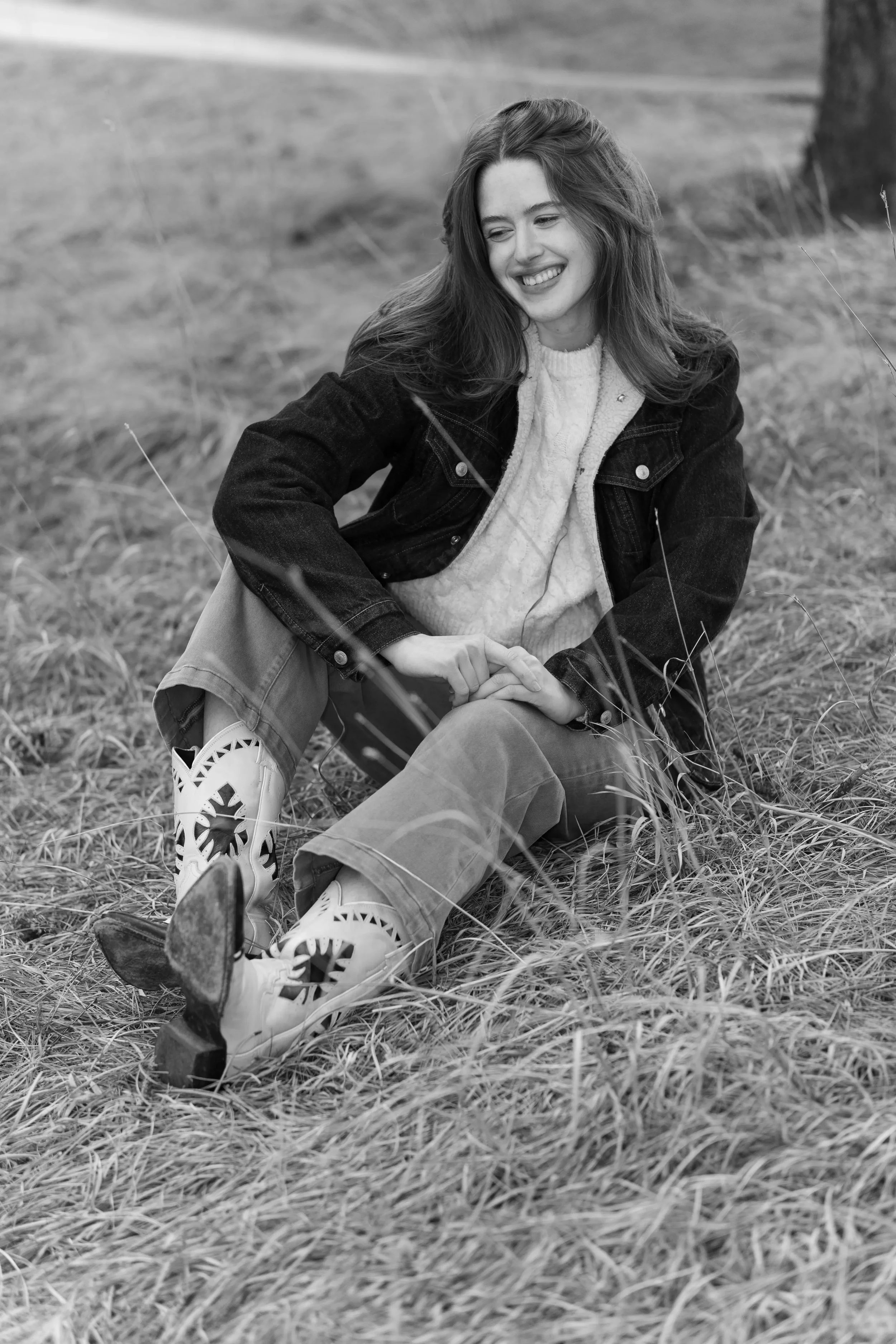 A young woman with long hair sitting on the grass outdoors, smiling with her eyes closed, wearing a dark jacket, light-colored sweater, light pants, and sneakers.