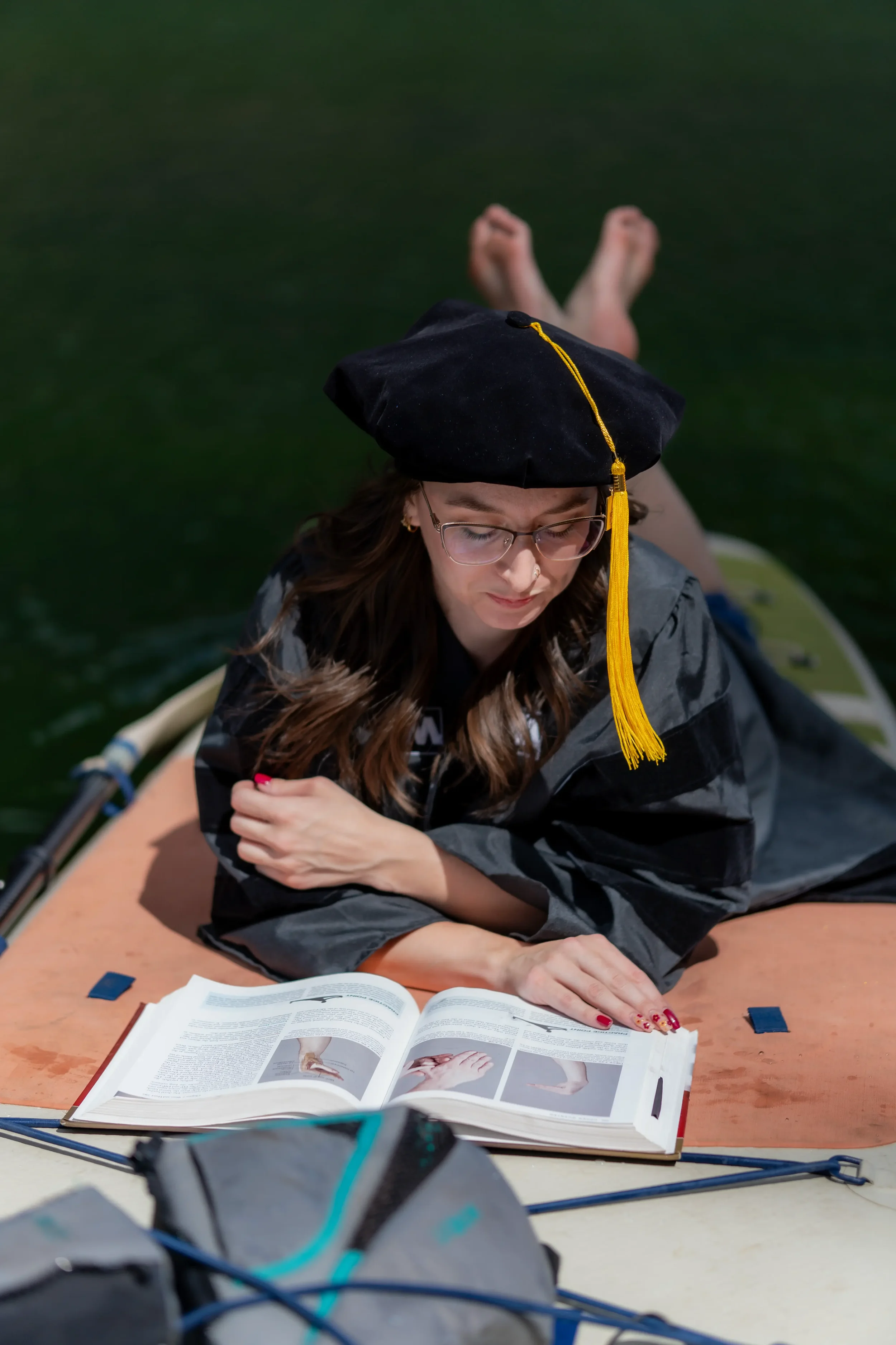Graduate wearing a black cap and gown, glasses, and earrings, lying on her stomach reading a book on a table in an outdoor setting with water in the background.