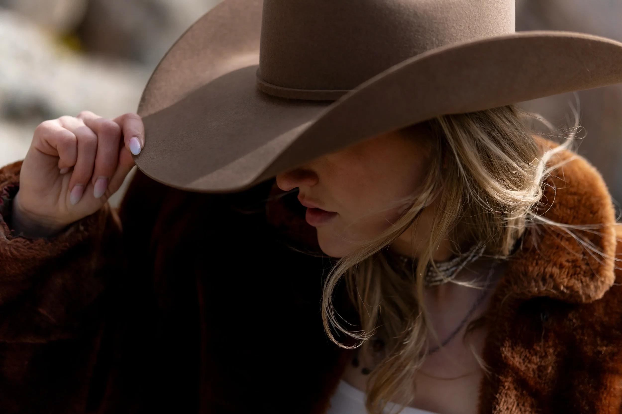 A woman with blonde hair wearing a brown felt hat and a fuzzy brown jacket, holding her hat with her left hand.