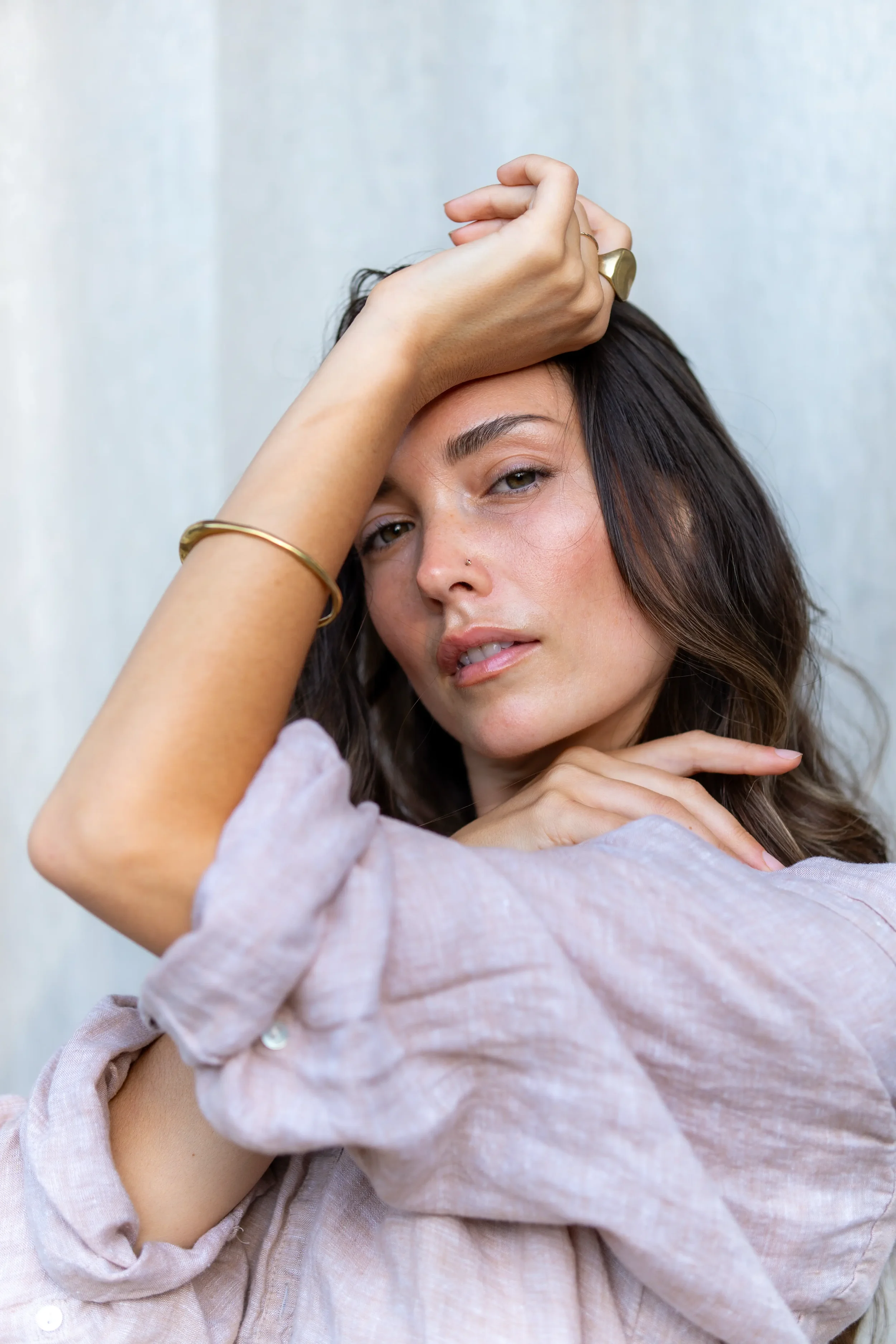 A woman with brown hair wearing a light pink shirt and gold jewelry, resting her head on her crossed arms, with one hand on her forehead and the other touching her chin, looking into the camera.