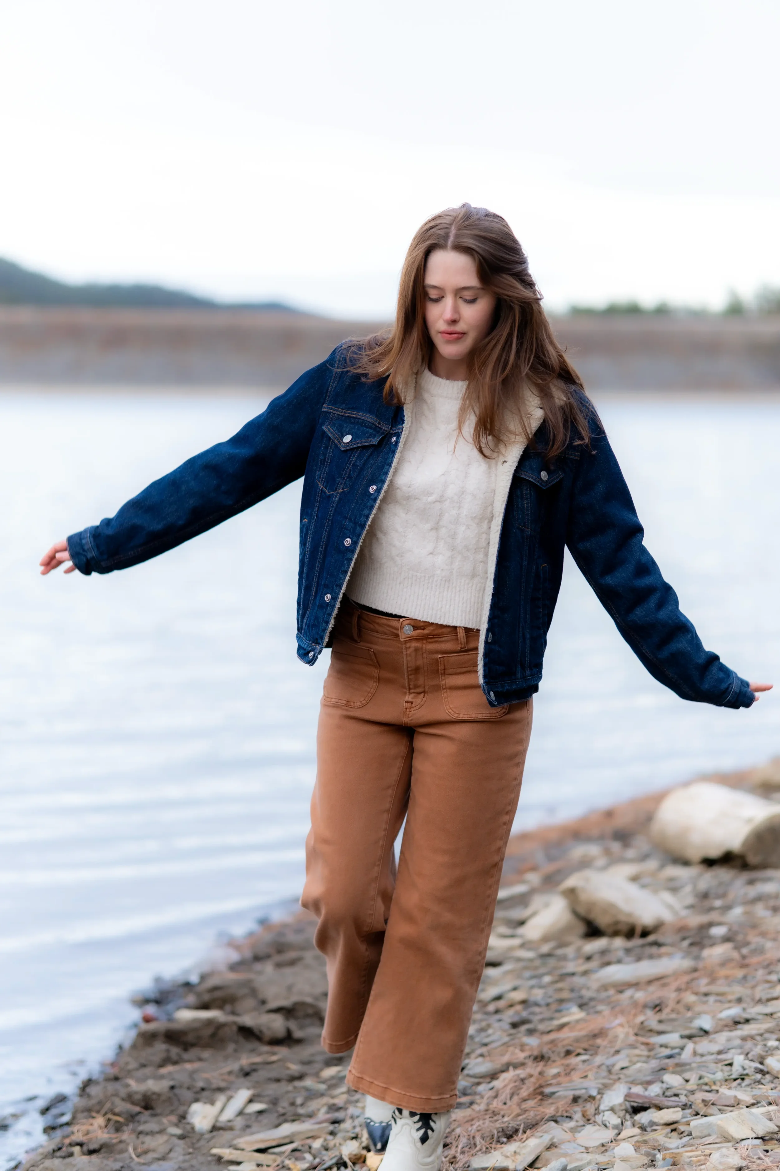Young woman with long brown hair standing on rocky shoreline near water, wearing a denim jacket over a cream sweater and brown pants, looking down with her arms outstretched.