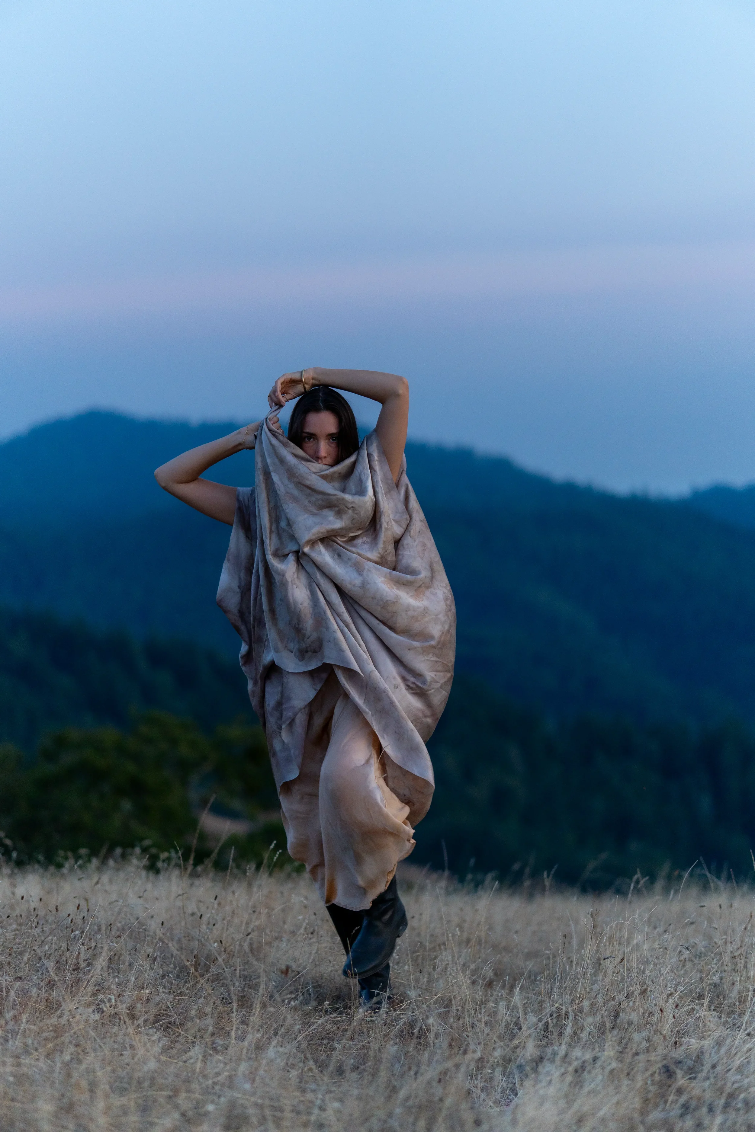 A woman standing in a field with mountains in the background, wearing a loosely wrapped cloth and black boots, with her arm raised and partly covering her face, during dusk or early evening.