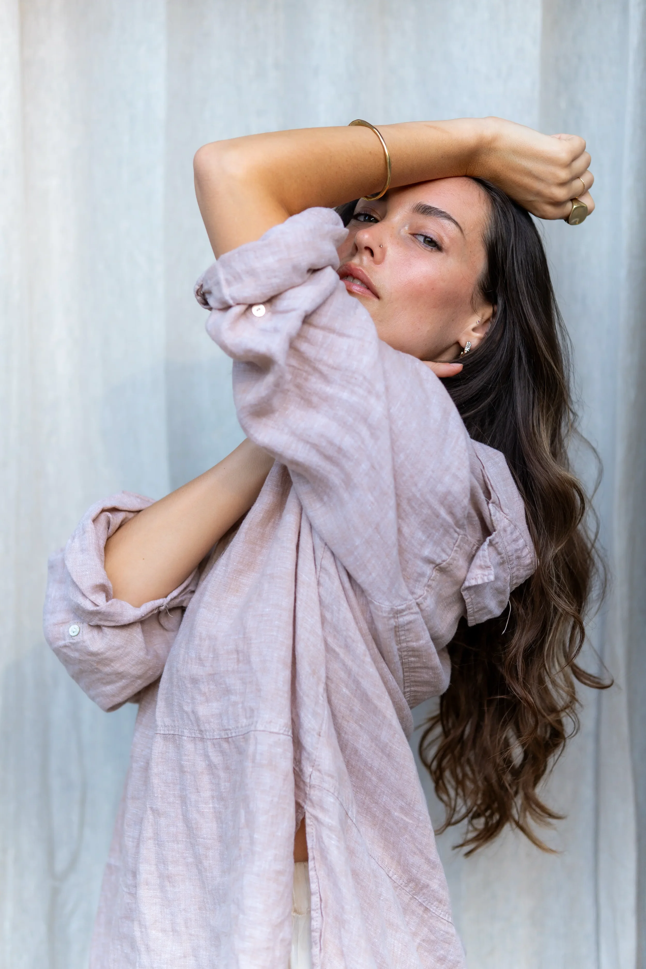 A woman with long wavy hair, wearing a light pink, slightly wrinkled blouse and jewelry, posing with her left arm raised and resting on her forehead against a neutral textured background.