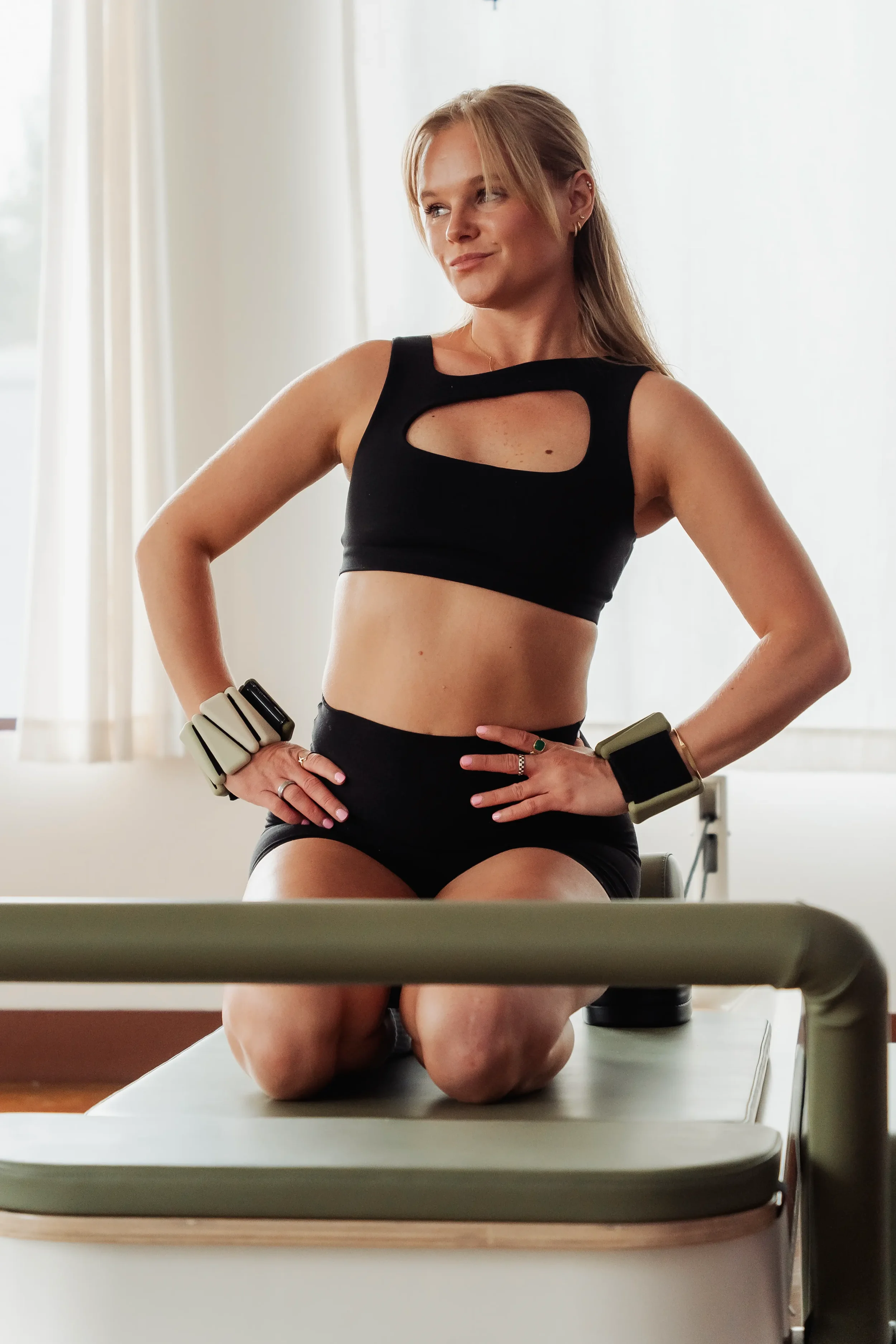 A woman wearing black workout clothes with cutouts, kneeling on an exercise table in a gym, with her hands on her hips, and looking to the side with a confident smile.