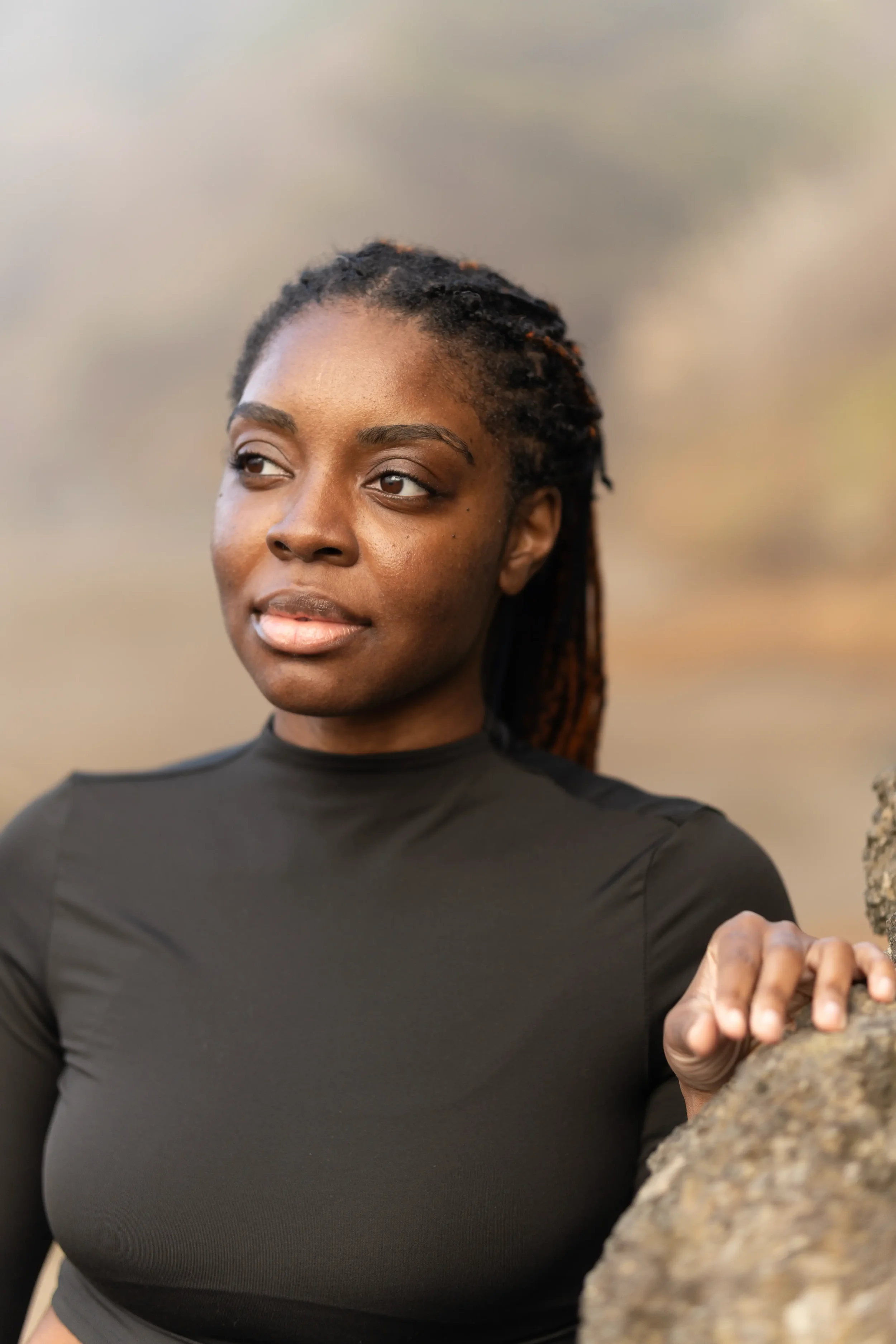 A woman with dark braided hair and a black long-sleeved shirt standing outdoors against a blurred background of trees and sky.