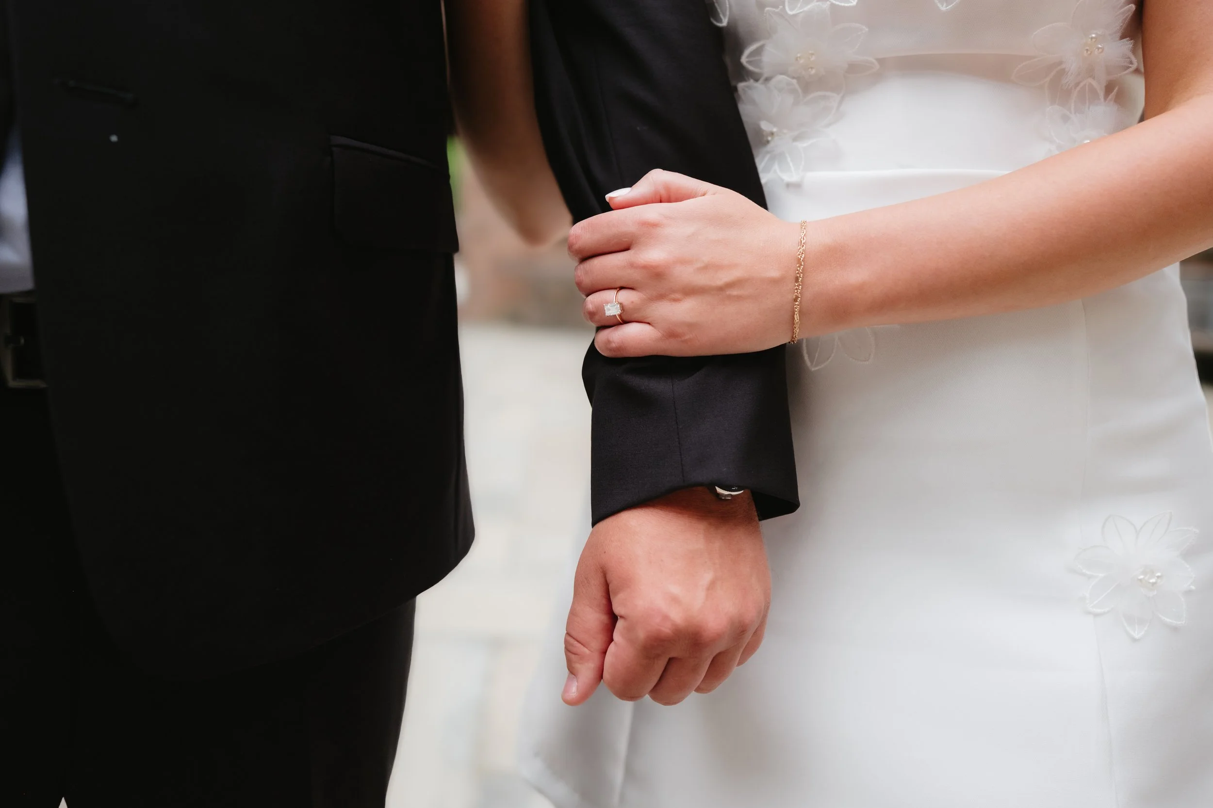 Bride in white dress with floral embroidery holding groom's arm, wearing a wedding ring.