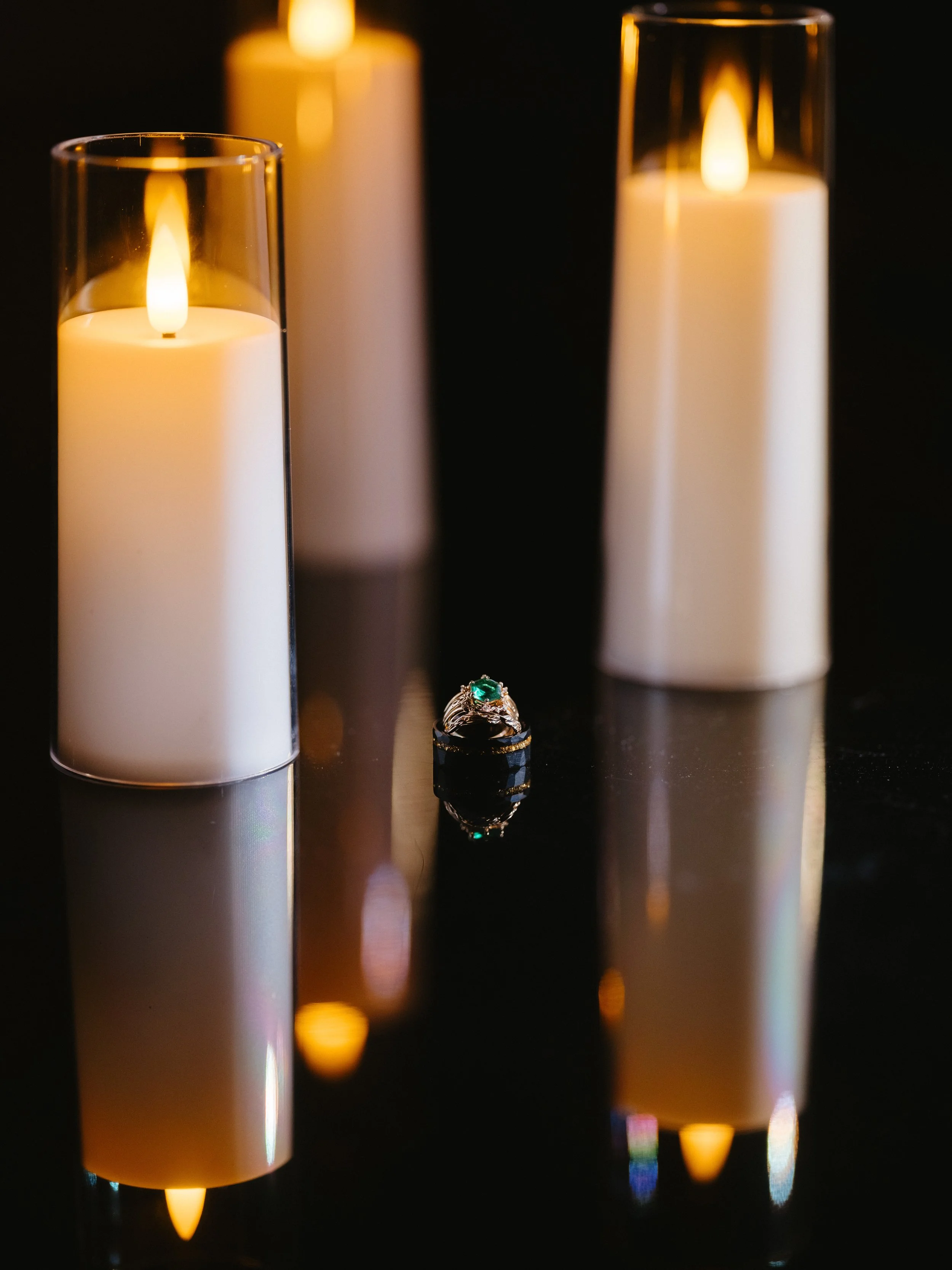 Three lit candles in glass holders and a ring with a green gemstone on a dark reflective surface.