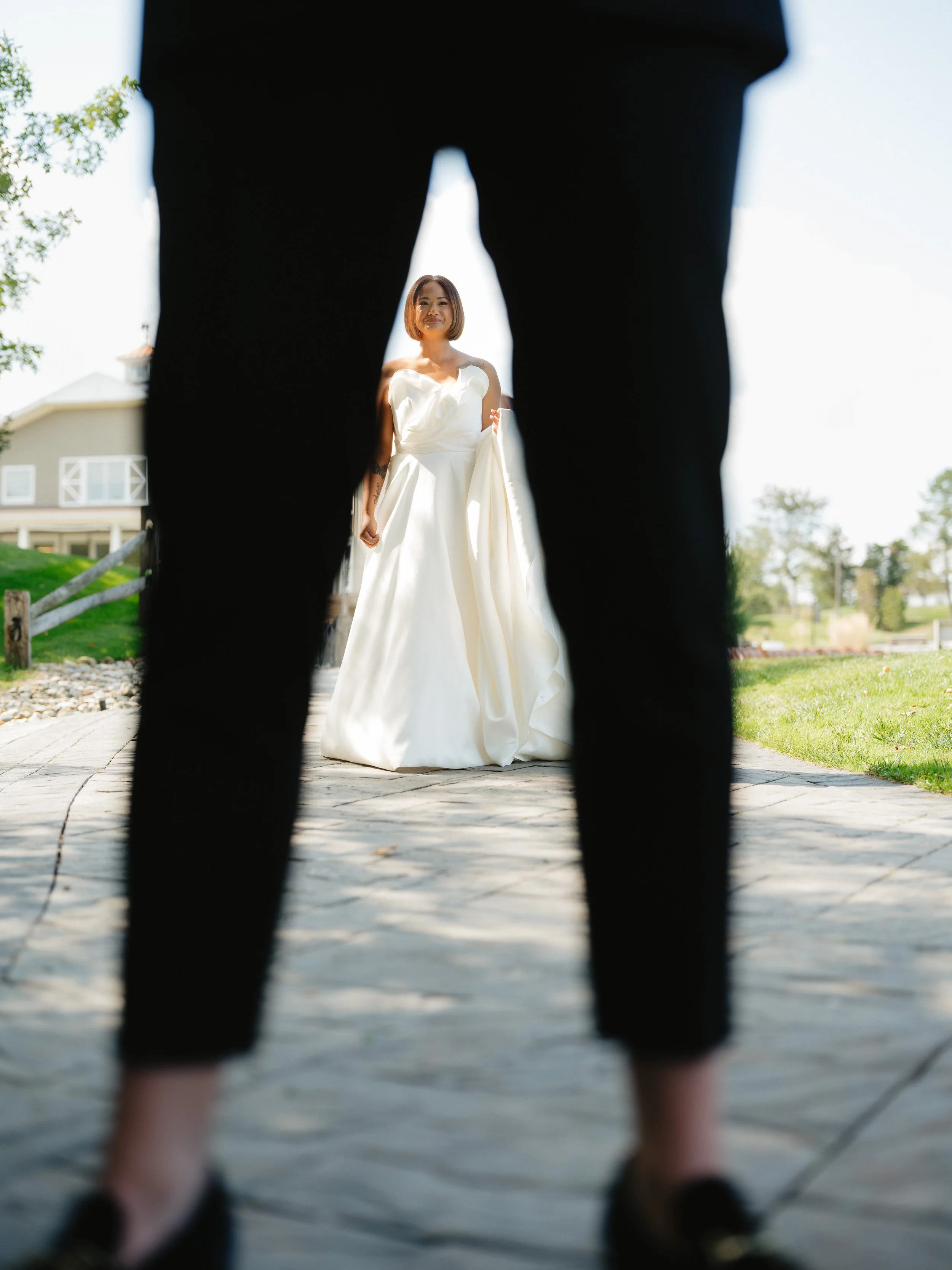 A bride in a white wedding dress is seen from between the legs of a person wearing black pants and shoes, with a garden and house in the background.