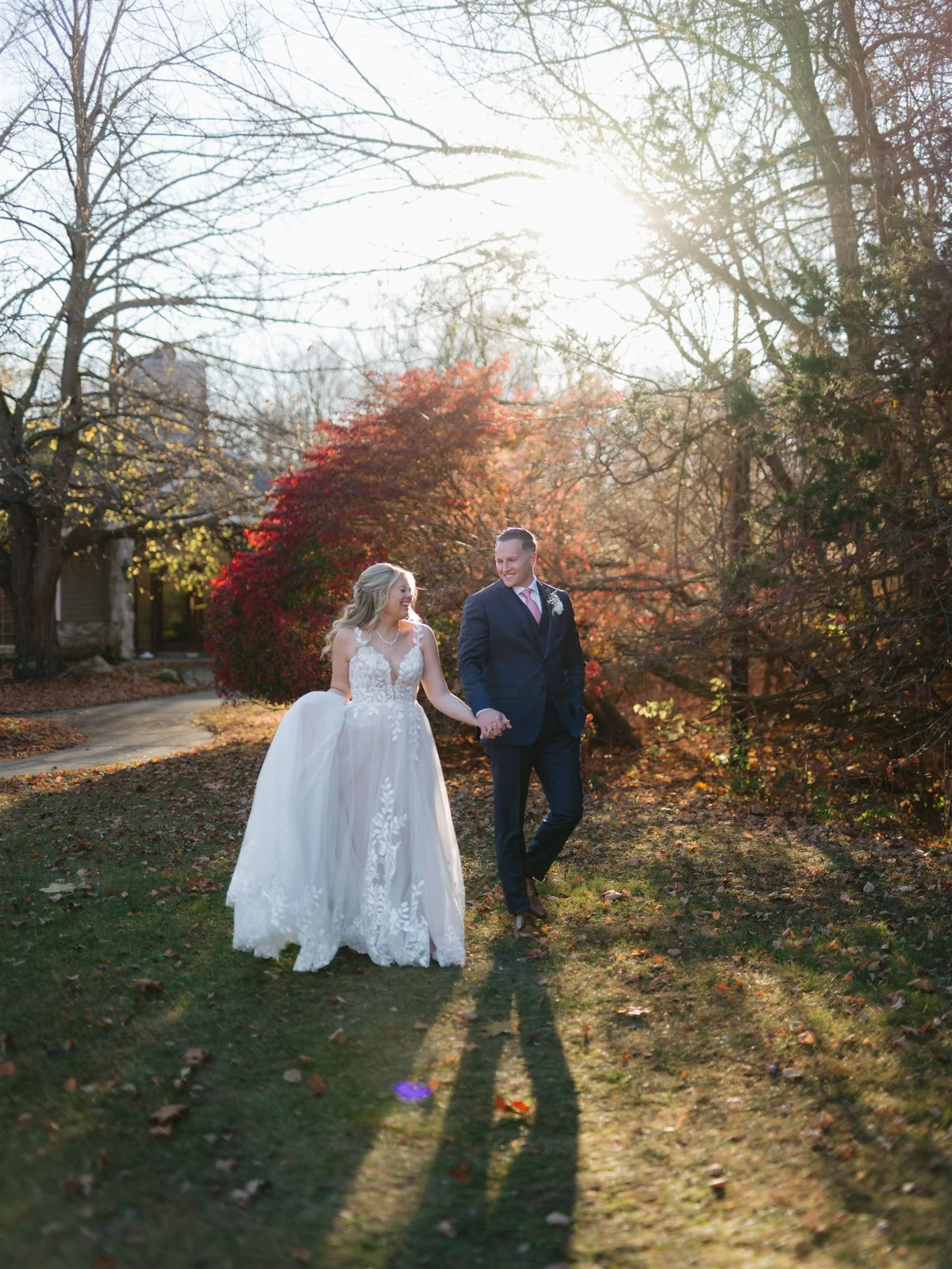 Bride and groom standing outdoors at sunset on a grassy field, holding hands and smiling at each other. The bride is wearing a white wedding dress with floral lace details, and the groom is in a dark suit with a boutonnière.