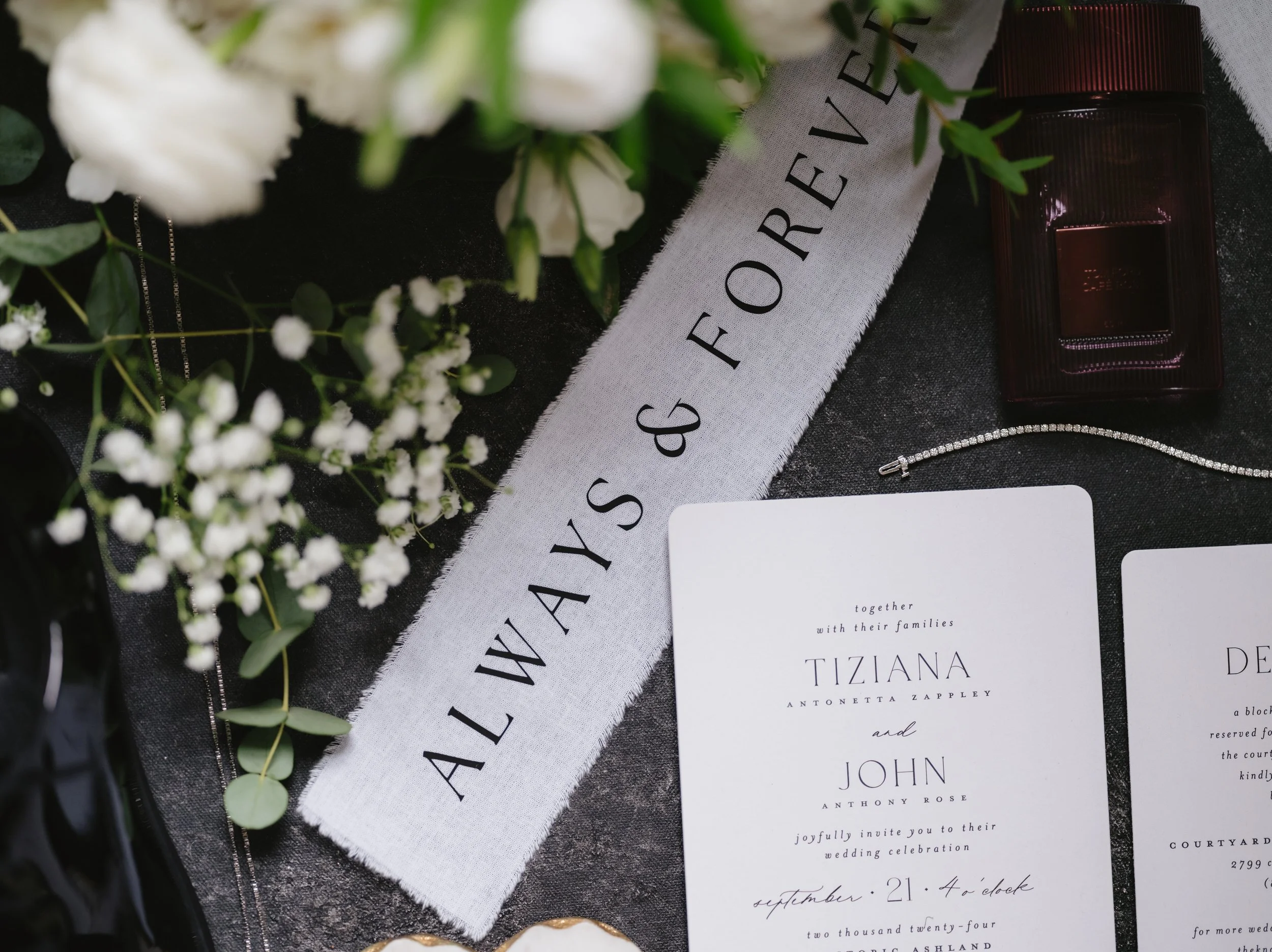 Wedding invitation flat lay with "Always & Forever" fabric, perfume bottle, white flowers, and jewelry on dark background.