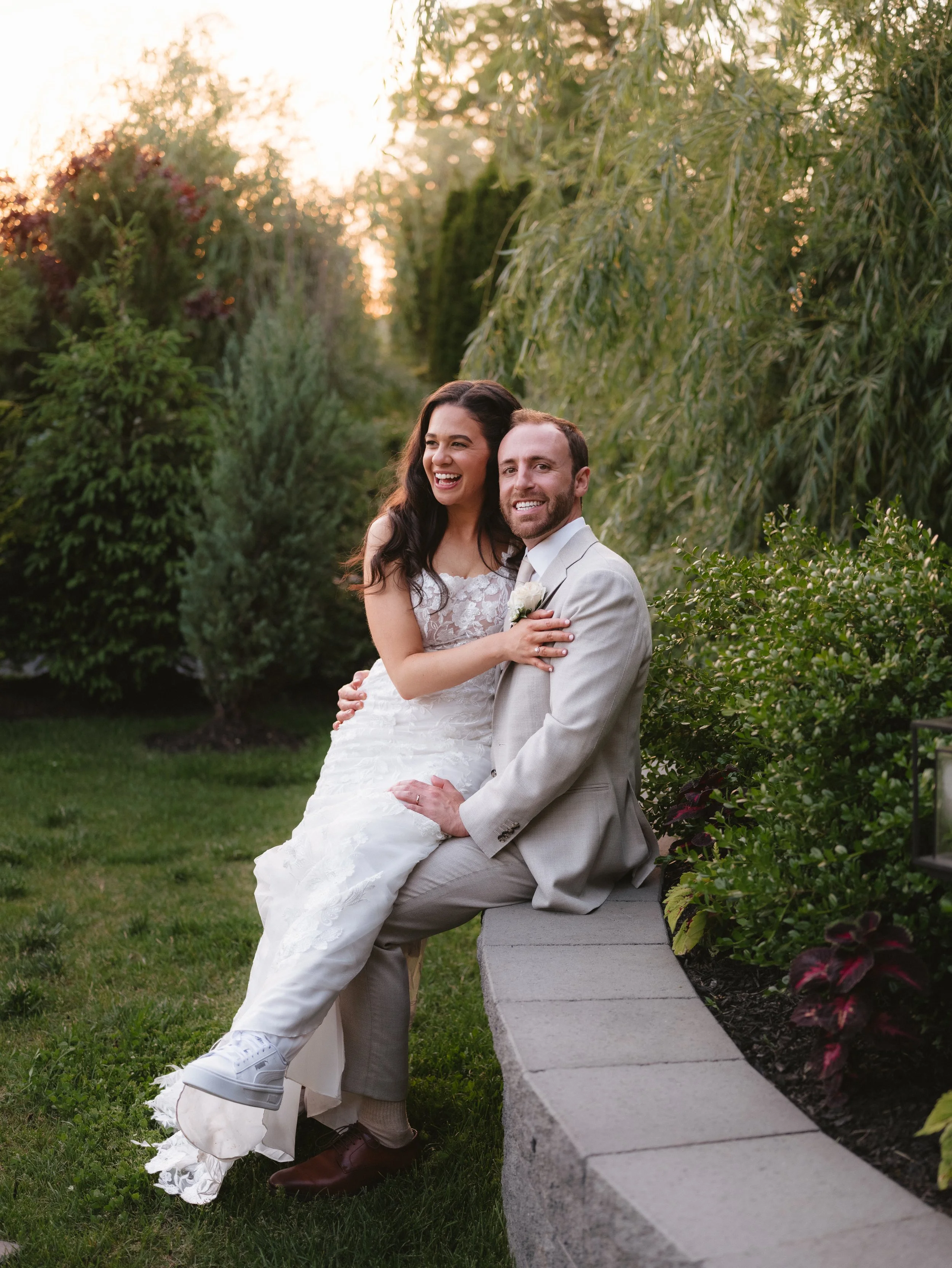 A bride and groom sitting on a stone wall, surrounded by greenery, with the bride in a white dress and sneakers, and the groom in a beige suit.
