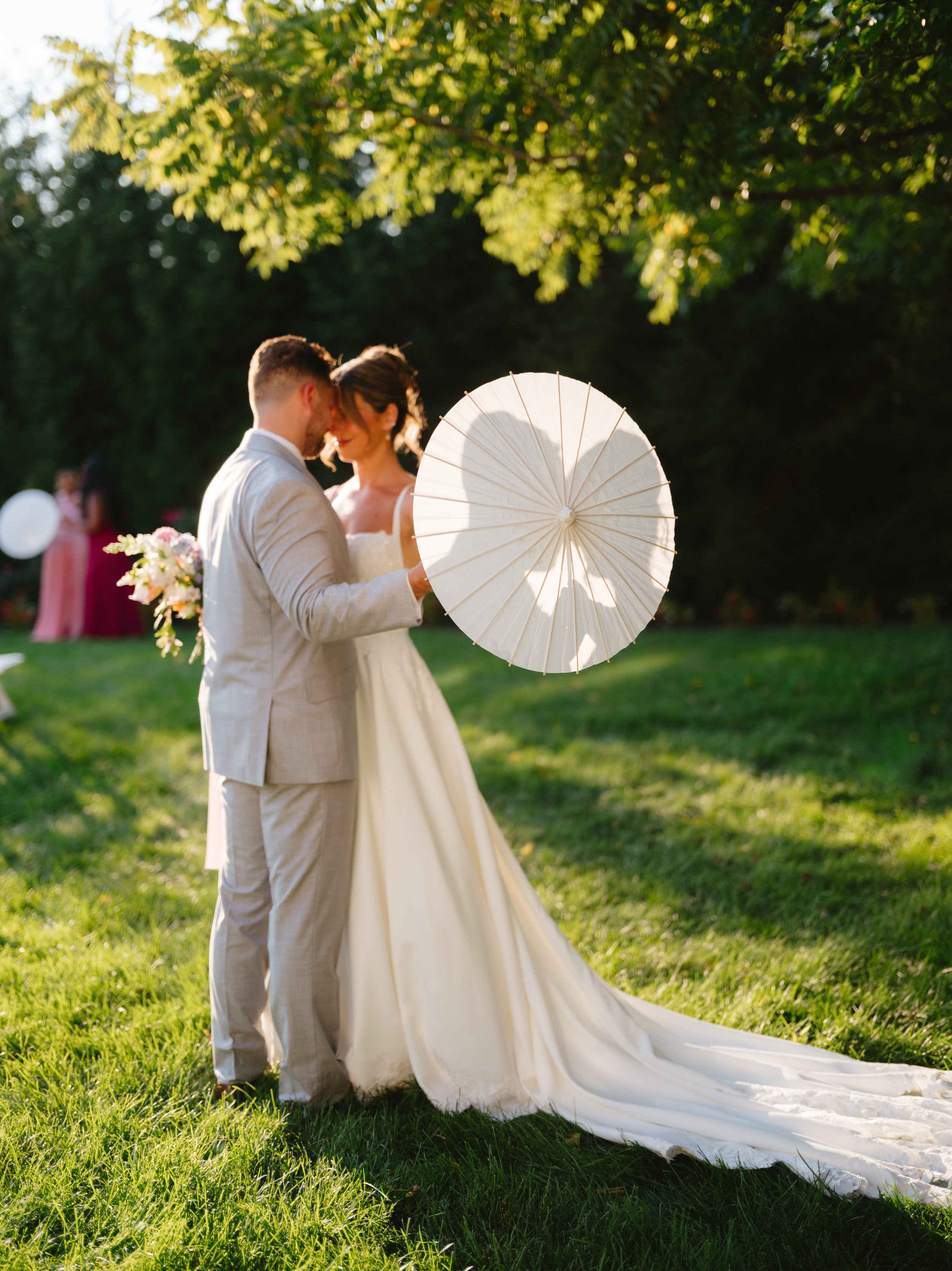 A bride and groom embrace outdoors during their wedding, holding a white parasol with the bride wearing a long white gown and the groom in a light-colored suit, surrounded by green trees and sunlight.