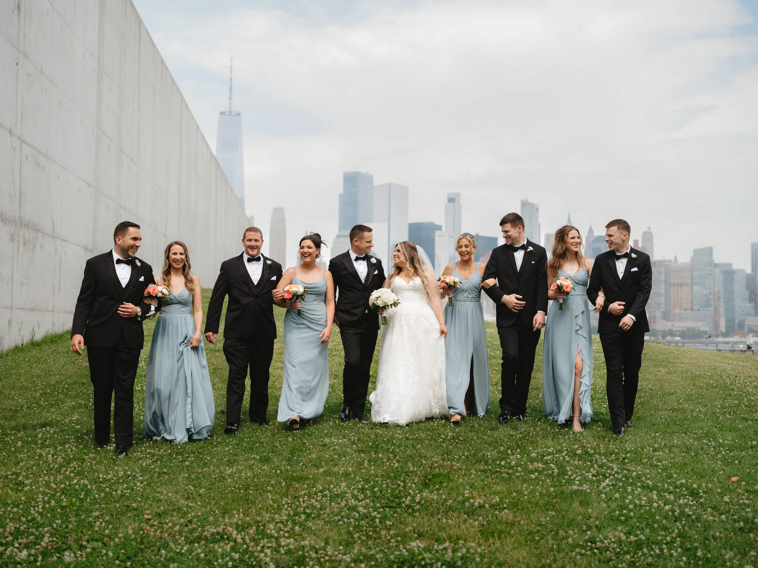 A wedding party walks on a grassy area near a concrete wall, with a city skyline in the background. The group consists of three men in black tuxedos and five women in blue dresses, along with one woman in a white wedding gown. They are holding bouquets and appear to be celebrating.