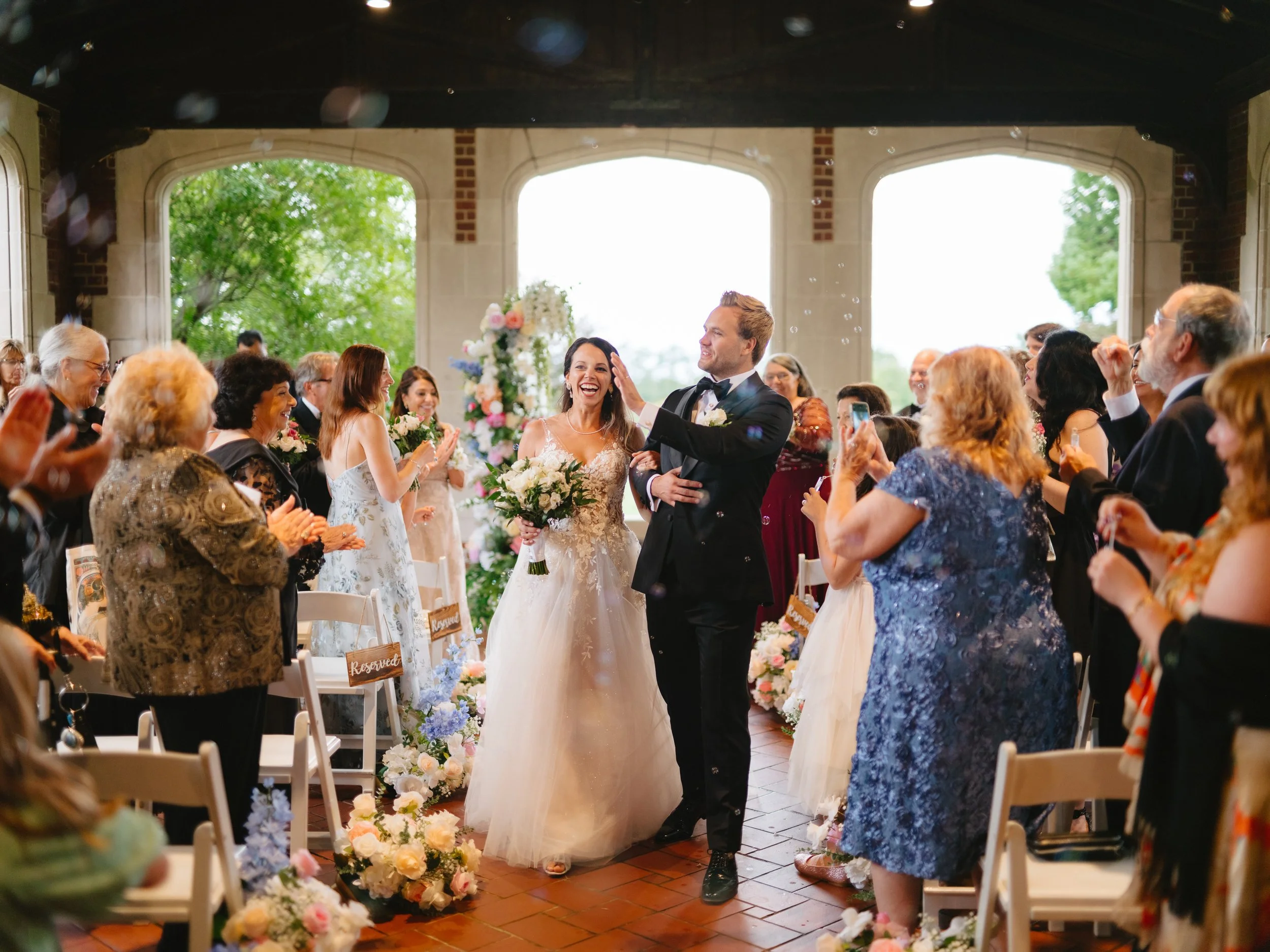 Bride and groom walking down the aisle with guests applauding in a decorated indoor venue.