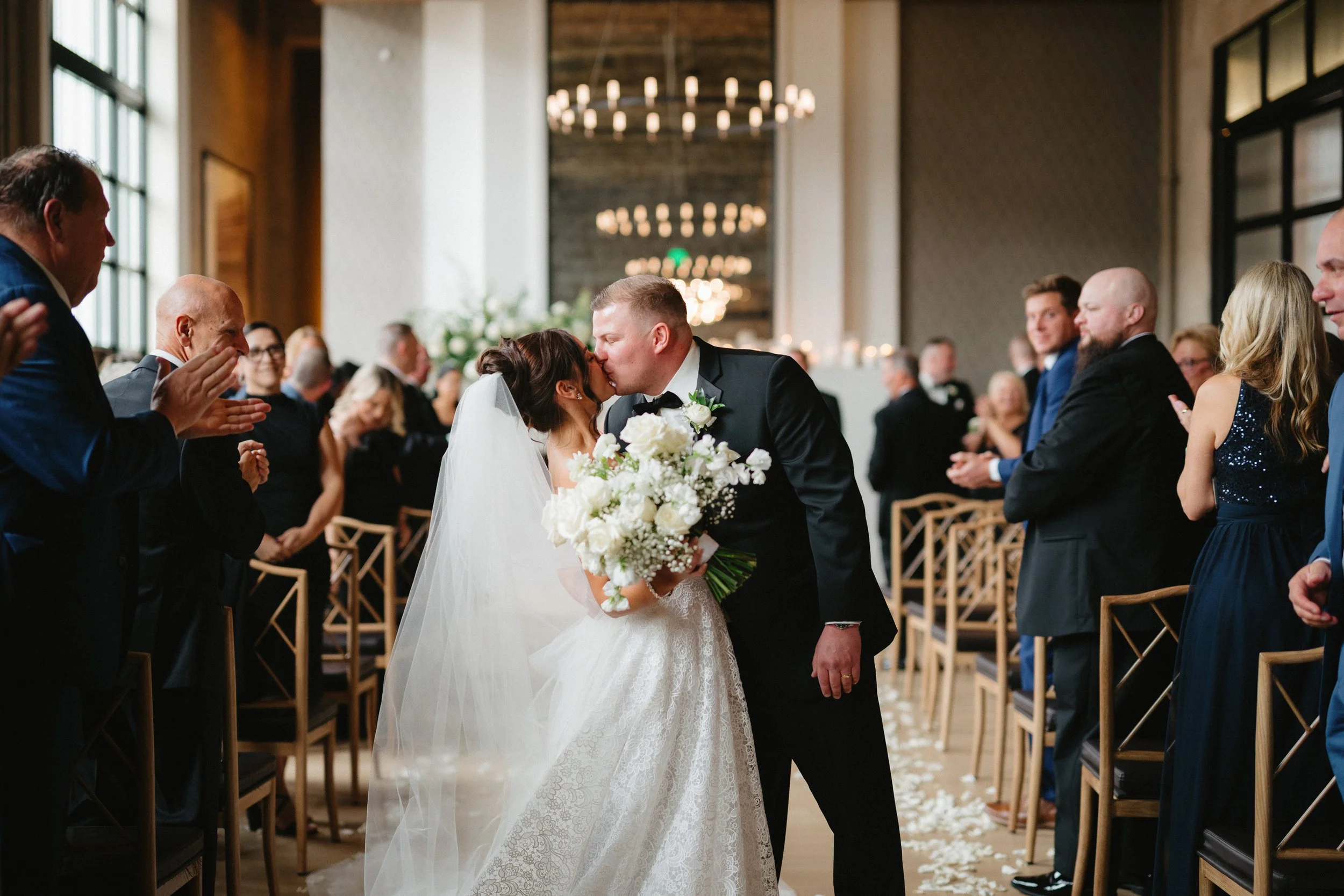Bride and groom kissing at the end of an indoor wedding aisle with guests applauding.