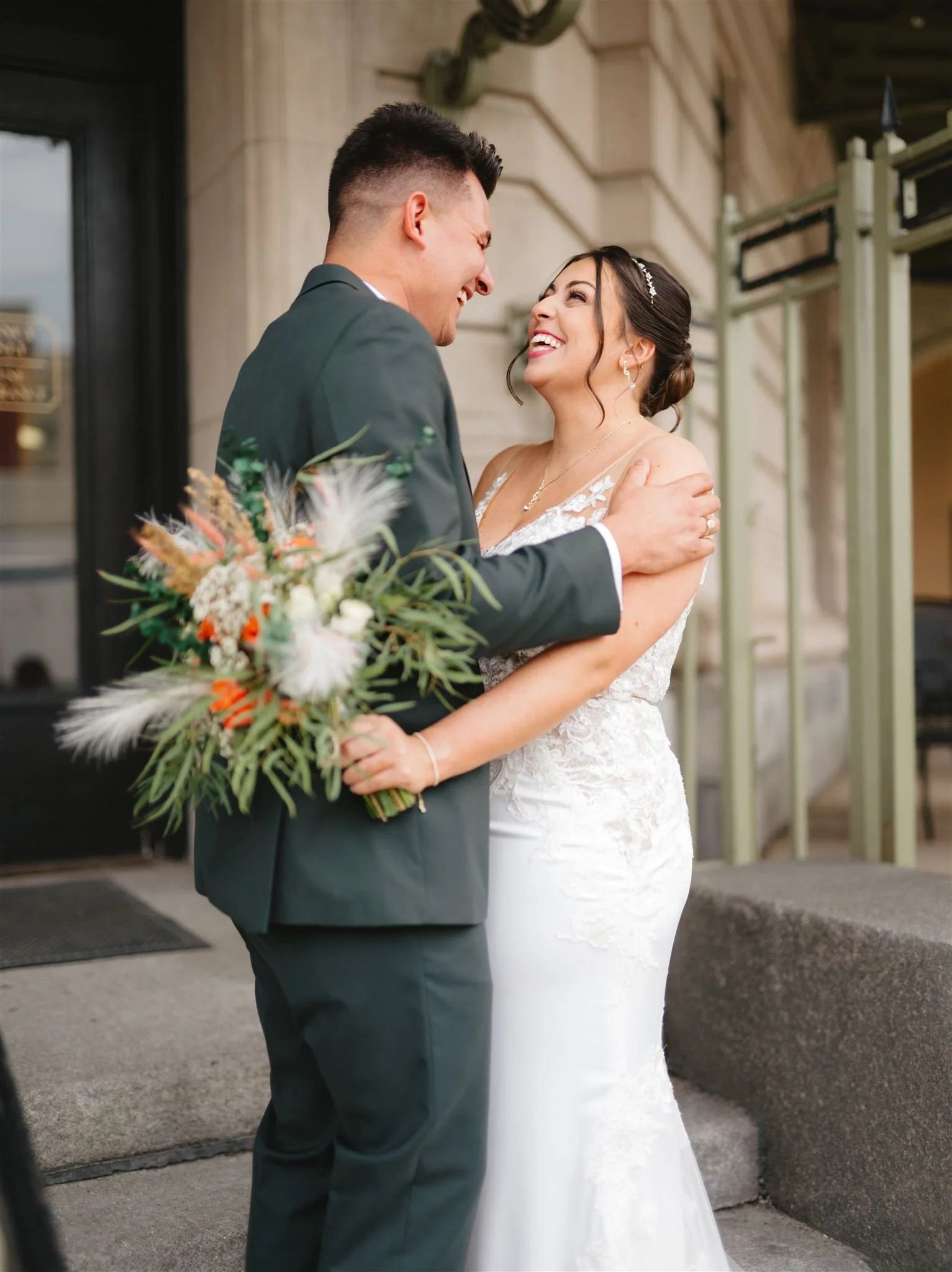 A couple in wedding attire smiling at each other; the man holds a bouquet.