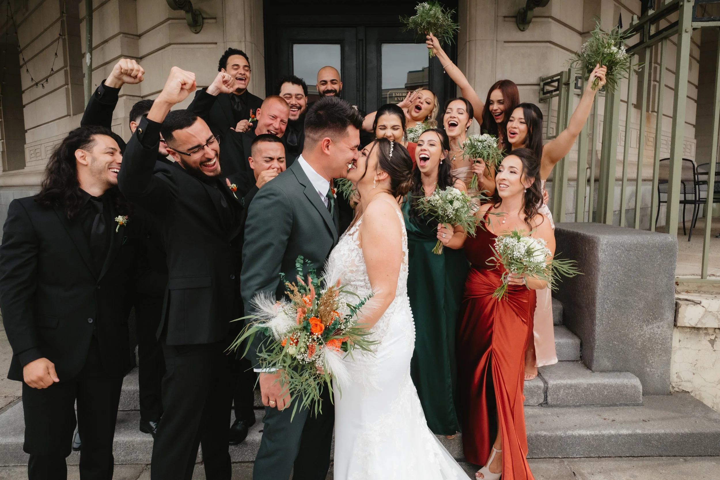 Wedding party celebrating on steps, bride and groom kissing, bridesmaids holding bouquets, groomsmen cheering.
