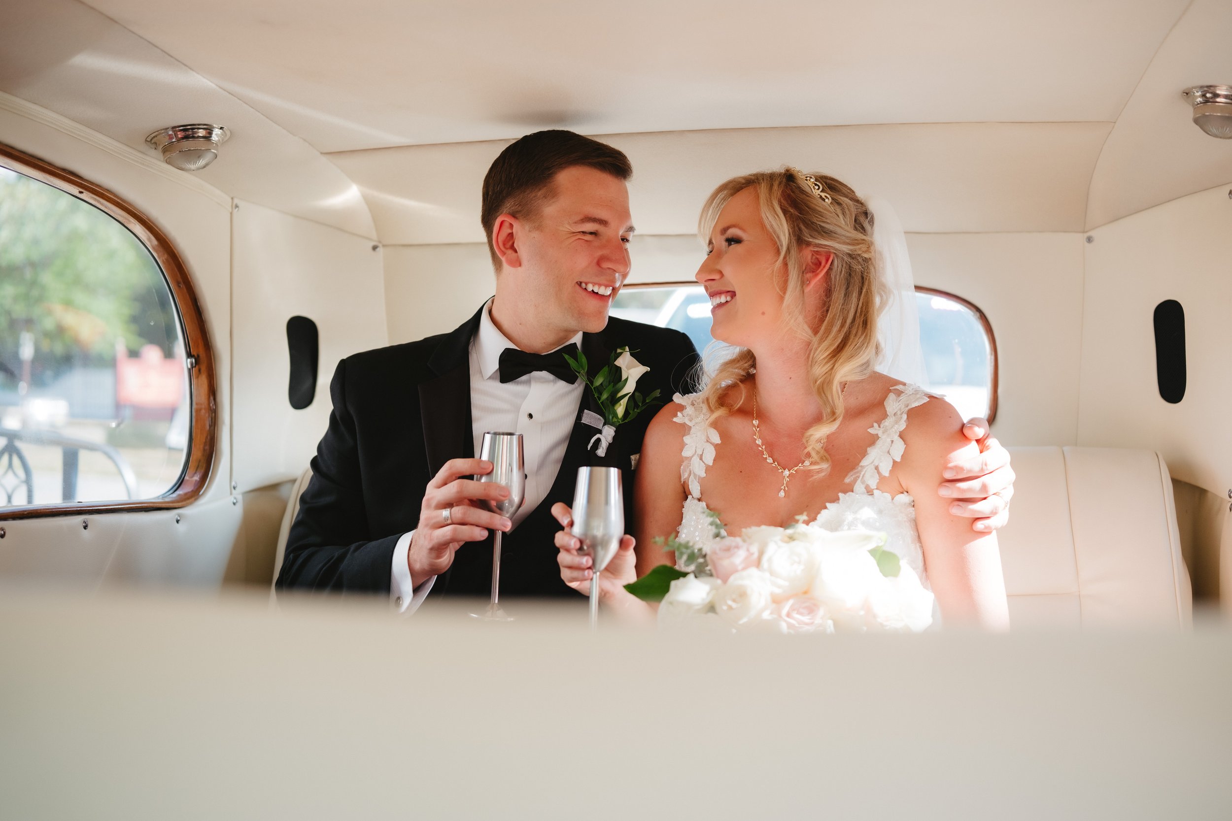Bride and groom smiling while holding glasses inside a car