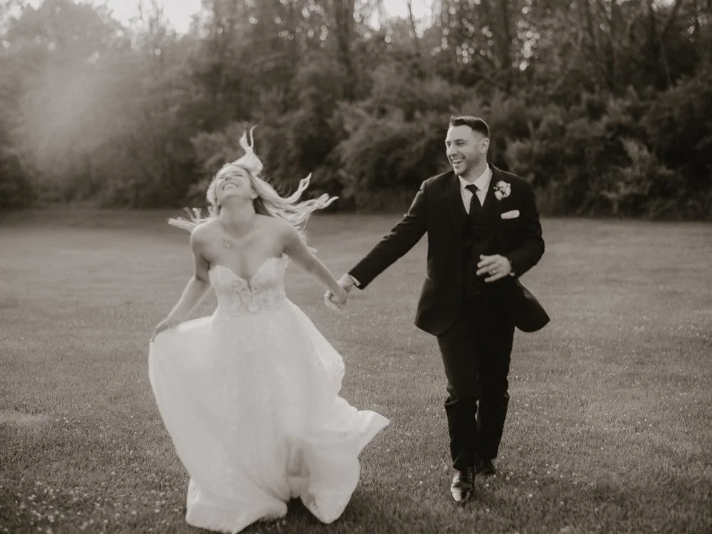 A bride and groom running joyfully across a grassy field. The bride is wearing a flowing white dress, and the groom is in a dark suit with a boutonniere, holding hands.